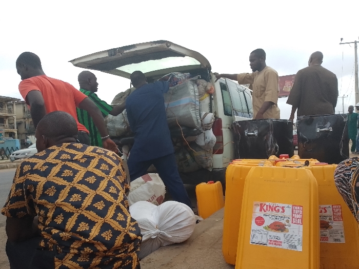 The scene depicts an outdoor commercial activity on a paved roadside in Mandate Estate, Ilorin, Nigeria. A white van with its rear tailgate open is centrally positioned, heavily loaded with numerous gray and white sacks. Six adult males are visible. Three individuals are actively manipulating cargo at the rear of the van: one in an orange t-shirt, one in a green and black striped shirt, and one in a dark blue tunic. A fourth male in a light brown traditional garment stands to the right of the van, observing. A fifth male in a dark brown traditional outfit is partially obscured behind the light brown-clad individual. In the immediate foreground, a sixth male, wearing a dark blue and gold patterned shirt, is seated or kneeling with his back to the viewer, next to white sacks on the ground. To the right of the van, a stack of multiple large, yellow plastic jerrycans, many labeled "KING'S PURE VEGETABLE OIL," rests on the ground, alongside several large black bags with handles. Buildings line the background on the left, and utility poles are visible against an overcast sky in the upper right.