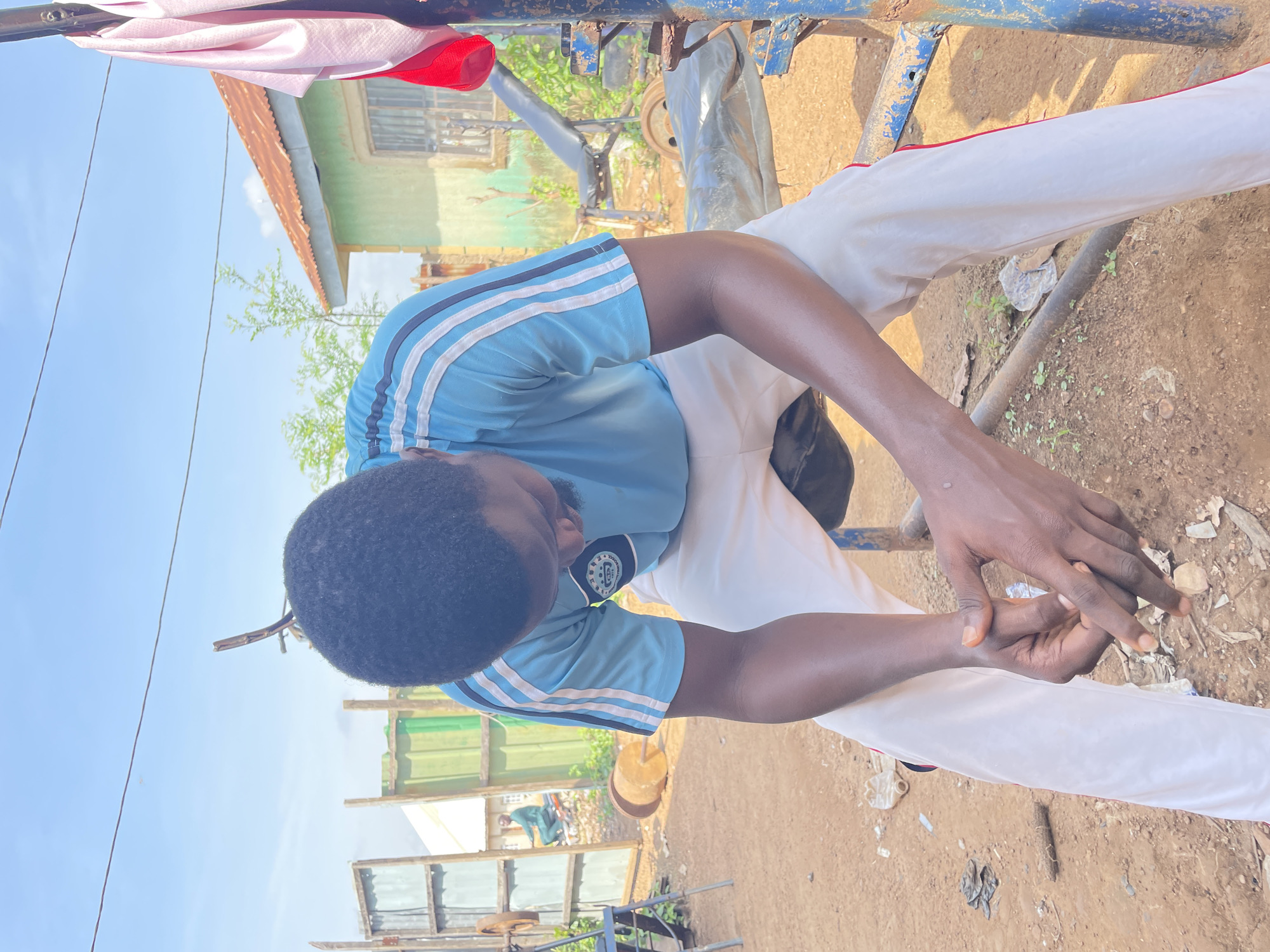 An individual is seated outdoors in Akwanga, Nigeria, in what appears to be a makeshift outdoor gym. The individual, wearing a blue t-shirt with white and black stripes and white pants with red trim, is looking down with their hands clasped. The surrounding environment is dusty and unpaved, with some visible debris. In the background, there are various objects that suggest a workout area, including a weight bench, weights, and metal framing. Structures resembling simple buildings and fencing are also present, along with some vegetation. The sky is clear and blue, indicating daylight.