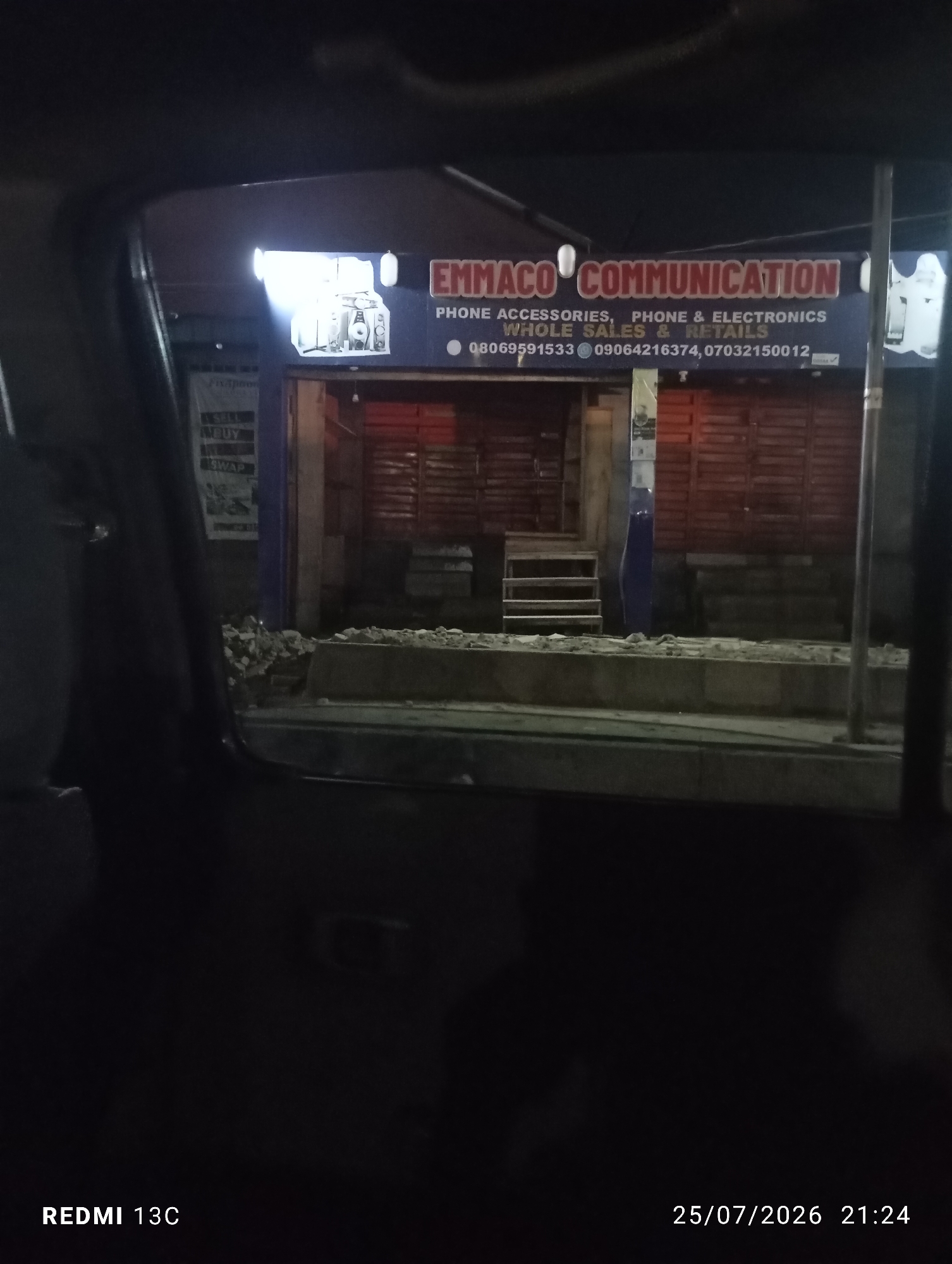 A nighttime, medium shot captures a storefront in Abeokuta, Nigeria, as viewed from inside a vehicle. The storefront, labeled "EMMACO COMMUNICATION," displays a sign indicating "PHONE ACCESSORIES, PHONE & ELECTRONICS, WHOLE SALES & RETAILS." Below this, contact numbers are listed: "08069591533, 09064216374, 07032150012." A banner on the left side of the entrance reads "SELL BUY SWAP". The store entrance is flanked by wooden shutters. Debris is visible on the sidewalk in front of the store, suggesting ongoing construction or repair work. The timestamp "25/07/2026 21:24" is present in the lower right corner, and "REDMI 13C" is in the lower left corner. The lighting is dim, with the storefront being the primary illuminated subject.