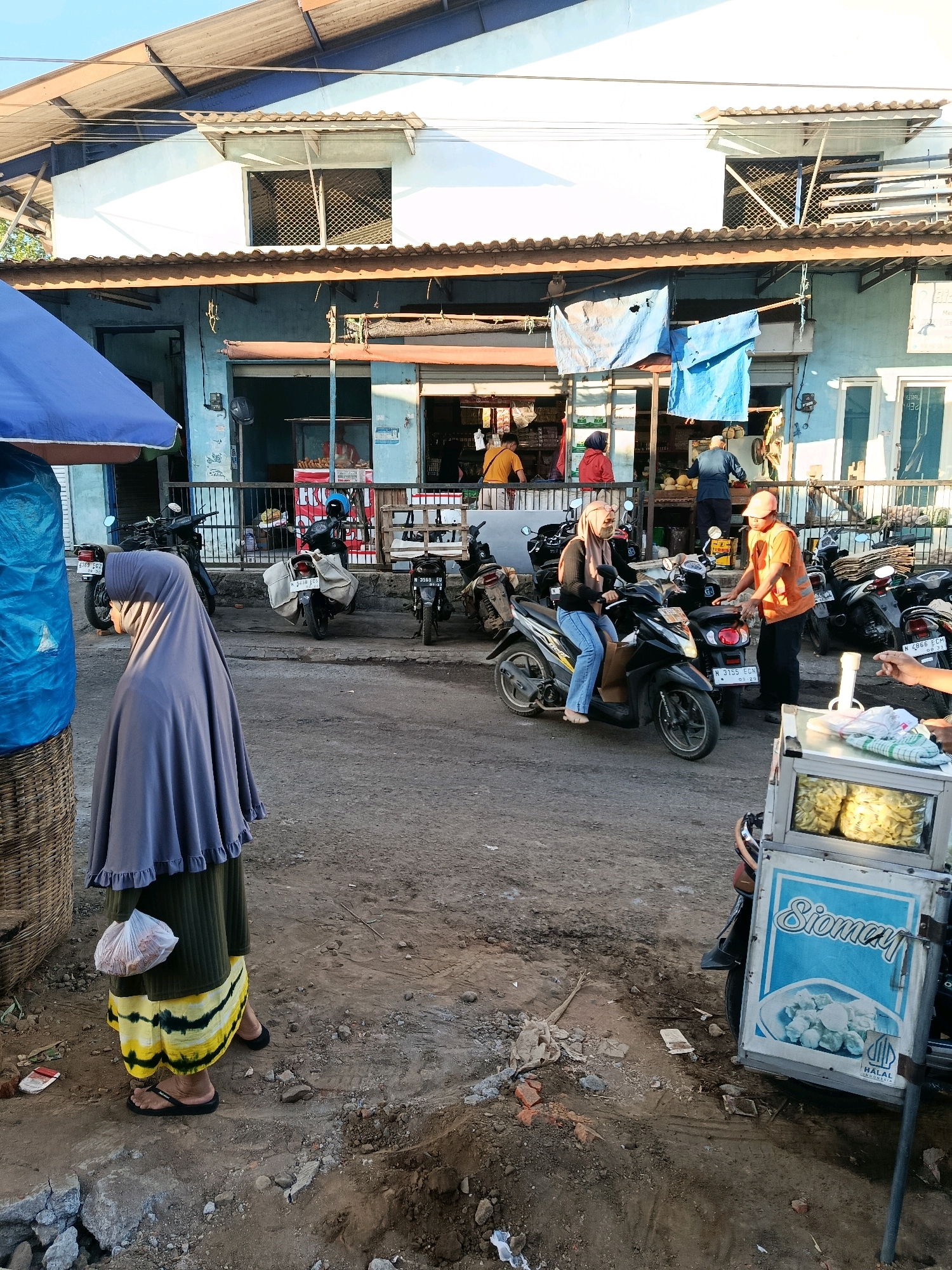 The image depicts a street scene in Bululawang, Indonesia, likely a market area. In the foreground, a woman wearing a gray hijab and a yellow and green patterned skirt stands facing away from the camera, holding a white plastic bag. To her left, a portion of a blue tarp and a woven basket are visible.
In the mid-ground, several motorcycles are parked. One black motorcycle has a woman wearing a beige hijab and a light-colored mask, riding it. A man in an orange vest and cap is standing next to her, interacting with her. Behind them, other motorcycles are parked, some with luggage.
Further back, a light blue building with a tiled roof forms the backdrop. There are shopfronts with open entrances. One shop appears to sell food items. In front of the shops, a mobile food cart labeled "Siomay" is visible, displaying a picture of the food and a "HALAL INDONESIA" logo. Various goods and items are displayed in the shop windows and on shelves. The ground is unpaved and appears to be dirt and gravel, with some debris. The lighting suggests it is daytime, possibly late afternoon, with sunlight casting shadows. The overall atmosphere suggests a busy, everyday commercial setting in a town.
