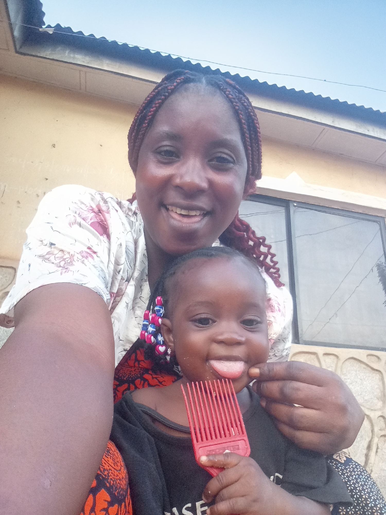 The image shows a woman and a young child in front of a building. The woman has braided hair with red highlights and is wearing a floral print top over an orange patterned skirt. She is smiling. The child is sticking out their tongue and holding a red comb. The child has decorations in their hair. The building behind them has a window and an exterior wall with a stone-like pattern. The location is Akwanga, Nigeria, based on the instructions.