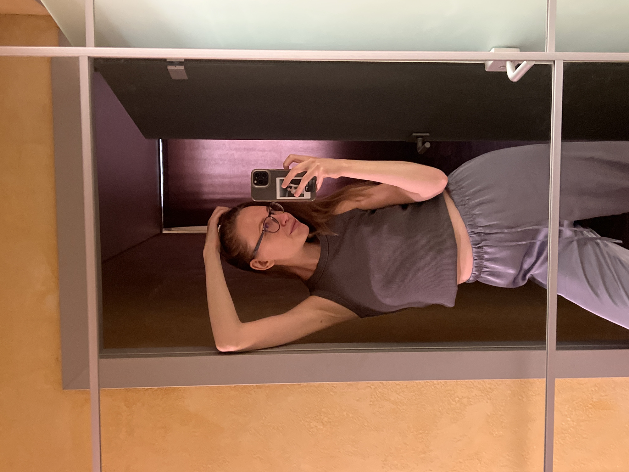 A woman is taking a selfie in a mirror. She is wearing glasses, a gray tank top, and gray pants. She is holding her phone in her right hand. The mirror is framed in gray and is hanging on a light-colored wall. A window with closed blinds is visible in the background. There is no visible location in Khimki, Russia.
