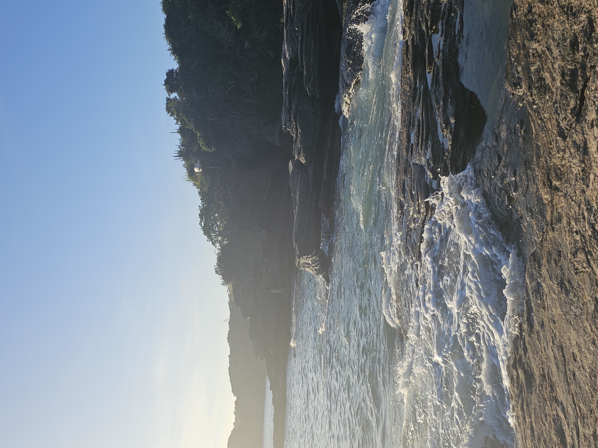 The image depicts a rocky coastline in Armação dos Búzios, Brazil. The sky is a clear blue, with a hazy, sunlit horizon. Lush green vegetation covers the cliffside. The ocean, churning with foamy white waves, crashes against the dark, jagged rocks. The scene is lit by bright sunlight.