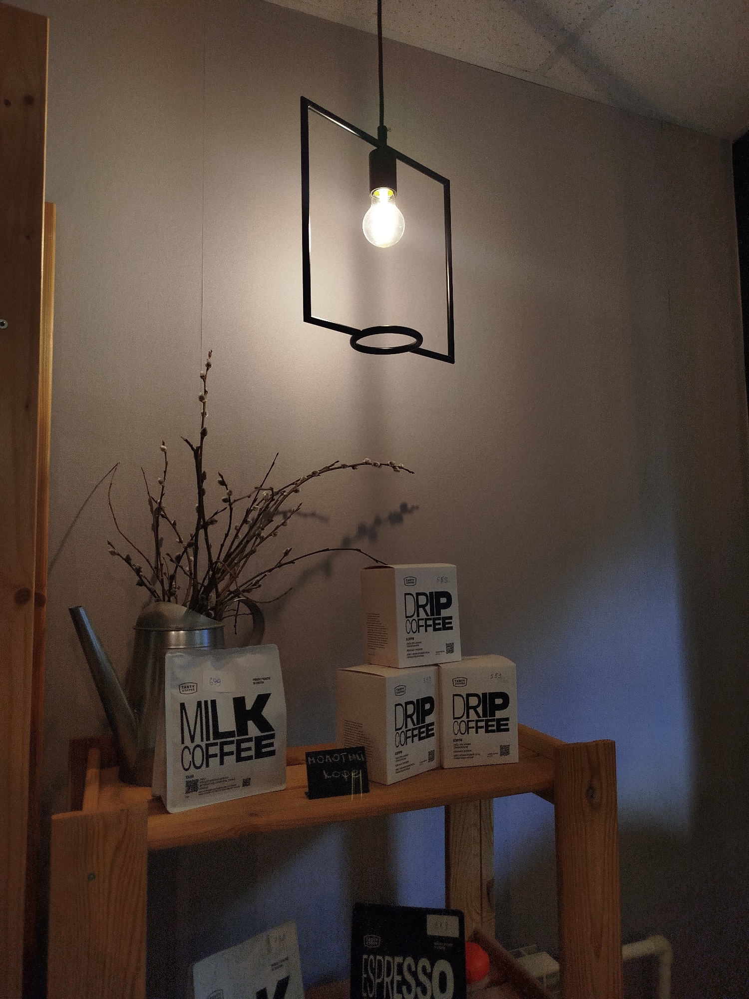 A still life scene is depicted indoors, featuring a wooden shelf displaying coffee-related items and decorative elements. The lighting is dim, with a single Edison-style bulb hanging from a minimalist black metal fixture above and casting shadows on the wall.
On the left side of the shelf, a vintage-style metal watering can holds a bouquet of bare branches with catkins. In front of the watering can, a silver bag labeled "MILK COFFEE" from "TASTY COFFEE" is prominently displayed. To the right of this bag, a small black chalk board displays handwritten text in a foreign language, appearing to be "МОЛОТИЛ" and "кофе".
To the right of the "MILK COFFEE" bag, three small cardboard boxes, stacked two high on the left and one on the right, are visible. Each box is labeled "DRIP COFFEE" from "TASTY COFFEE". The boxes are white with black text.
The background is a plain, textured wall, appearing to be a light gray or beige color. A wooden vertical beam is visible on the far left edge of the image, part of a larger structure, possibly a shelf unit or wall framing. The overall impression is one of a cozy, perhaps coffee-themed display in a domestic or commercial setting. There are no people or animals present in the image.