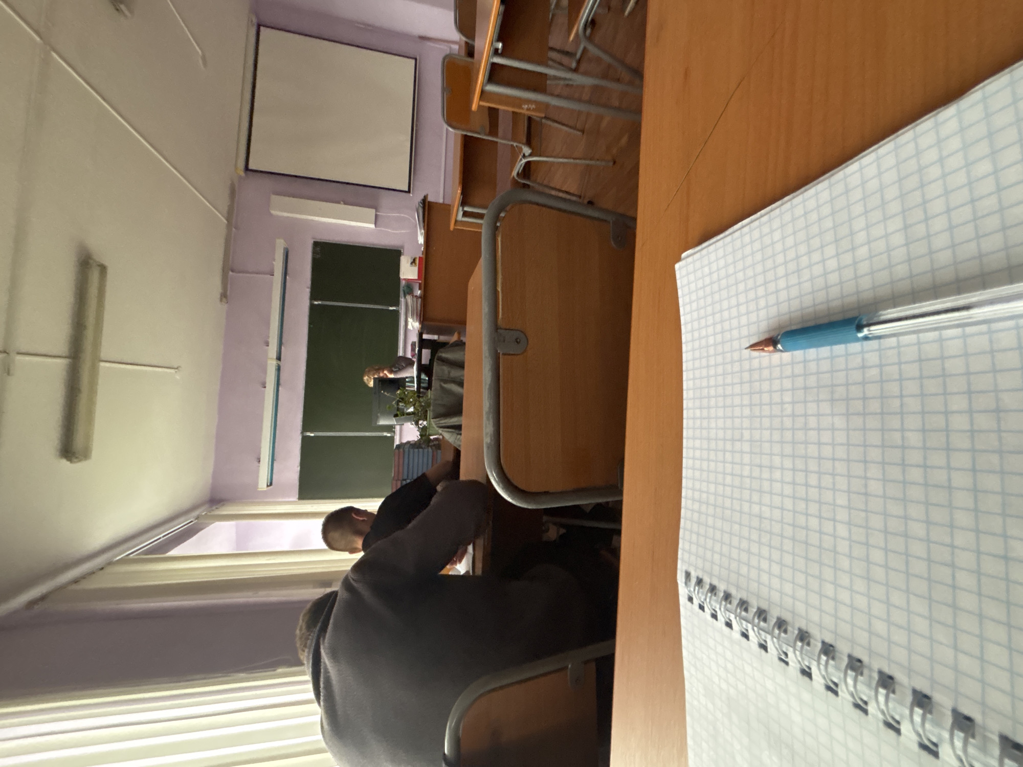 The image captures an interior view of a classroom or lecture hall, seen from a seated perspective. The scene is oriented vertically, implying a camera held sideways.
In the immediate foreground, a light brown wooden desk surface occupies the right side of the frame. An open, spiral-bound notebook with blue square-grid paper (graph paper) lies on this desk, and a blue and transparent ballpoint pen rests on its surface.
Moving further into the room, several rows of wooden desks with grey metal frames are visible. Two individuals are seated in the foreground, seen from behind. One person, wearing a dark grey hooded top, is seated closer to the viewer. Another person, wearing a dark shirt, is seated slightly further ahead. Both appear to be facing the front of the room. A third person is partially visible near the front of the classroom.
The front of the room features two large green chalkboards. Above them, a white projection screen is mounted on the wall. Below the chalkboards, there's a shelf or table holding a few potted plants and a stack of books or binders. The walls are a light purple color, and the ceiling is white with linear fluorescent light fixtures providing illumination. On the left side of the room, windows are covered by horizontal blinds.
The overall setting is that of an educational environment, likely during a class or lecture. Students are seated, and a presenter is at the front. No legible text is visible on the chalkboards, screen, or notebook. The time of day and weather conditions are not discernible from the image, and due to the perspective, no specific emotions can be observed.