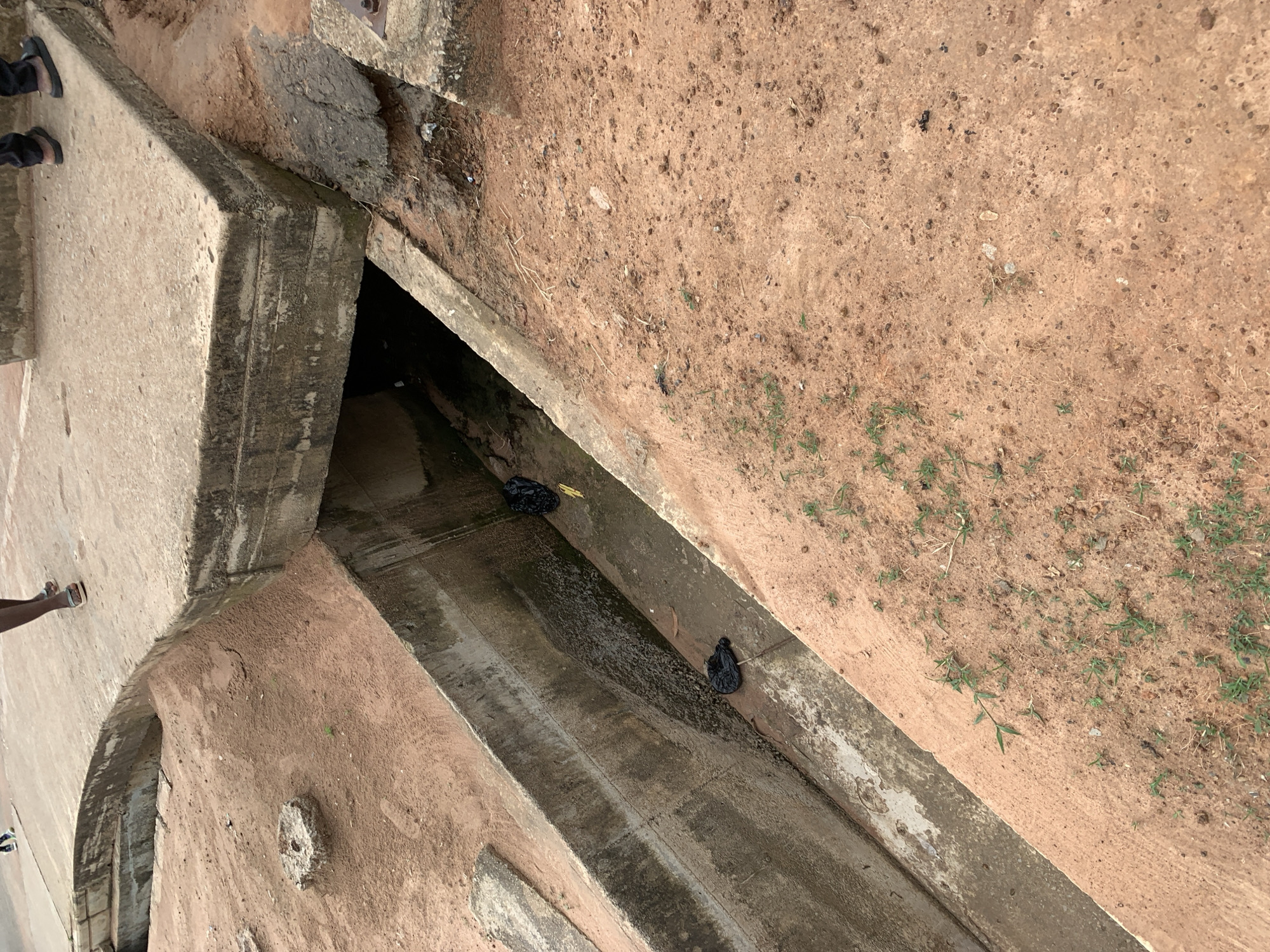 The image shows a street-level view of a drainage system in Wukari, Nigeria. The drainage channel is constructed of concrete and is partially filled with water and debris. There are several black plastic bags visible within the drainage channel, suggesting litter or waste. The surrounding area is covered in reddish-brown soil with sparse vegetation. A concrete walkway or structure is visible above the drainage channel, with at least two individuals standing on it, partially visible. One person is wearing black trousers and dark sandals, and the other person is wearing dark trousers and lighter sandals. The lighting suggests it is daytime.