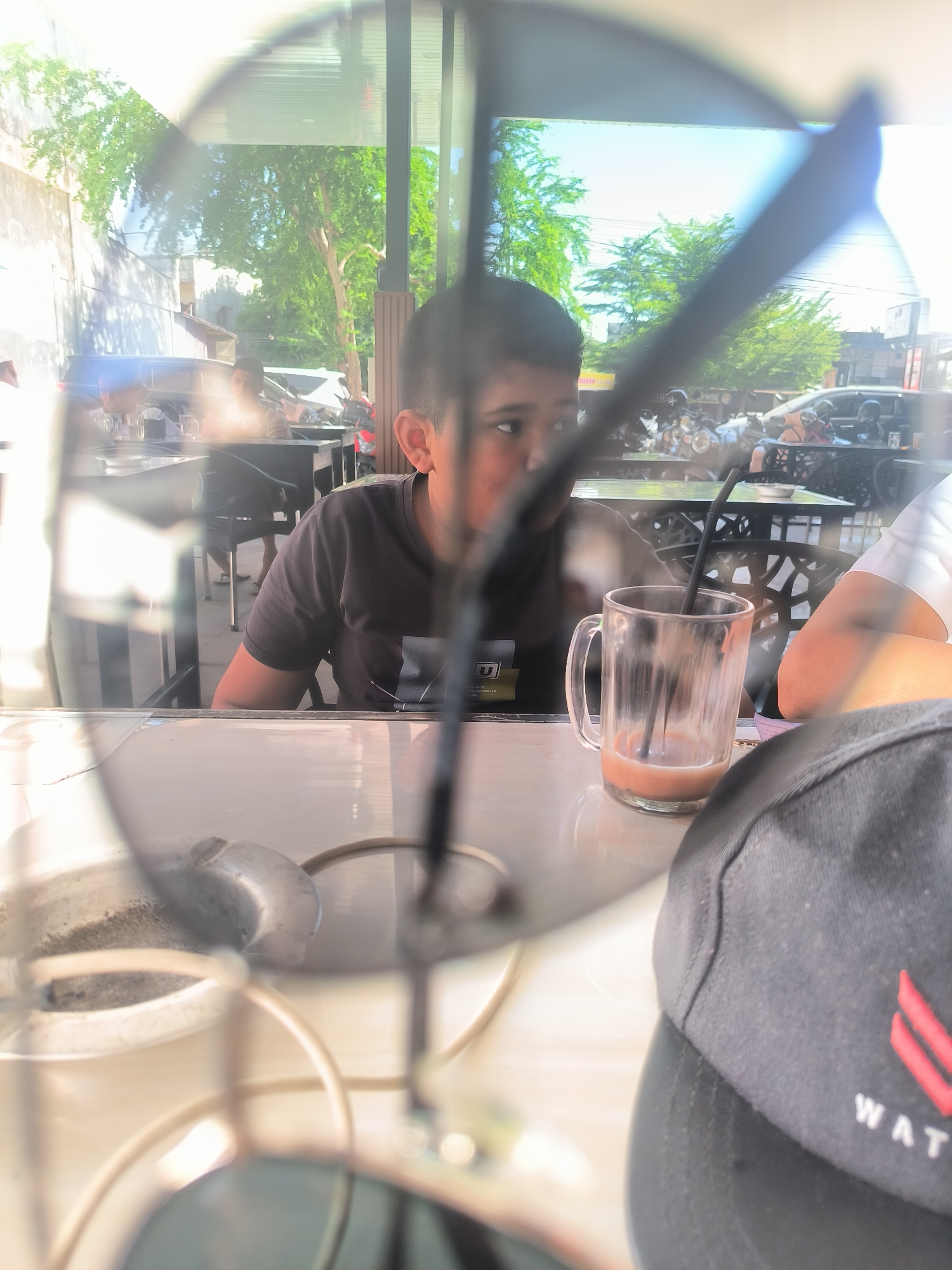 A boy is seated at a table in an outdoor café, possibly in Lhokseumawe, Indonesia. He is wearing a black t-shirt and looking to his right. On the table, there is a glass mug with a straw, and a black baseball cap with red and white lettering that reads "WAT" is visible in the foreground. Sunglasses are partially obscuring the foreground of the view. In the background, there are other tables and chairs with people seated at them, as well as motorcycles parked along the street. A building is visible in the background on the left, and trees can be seen in the distance.