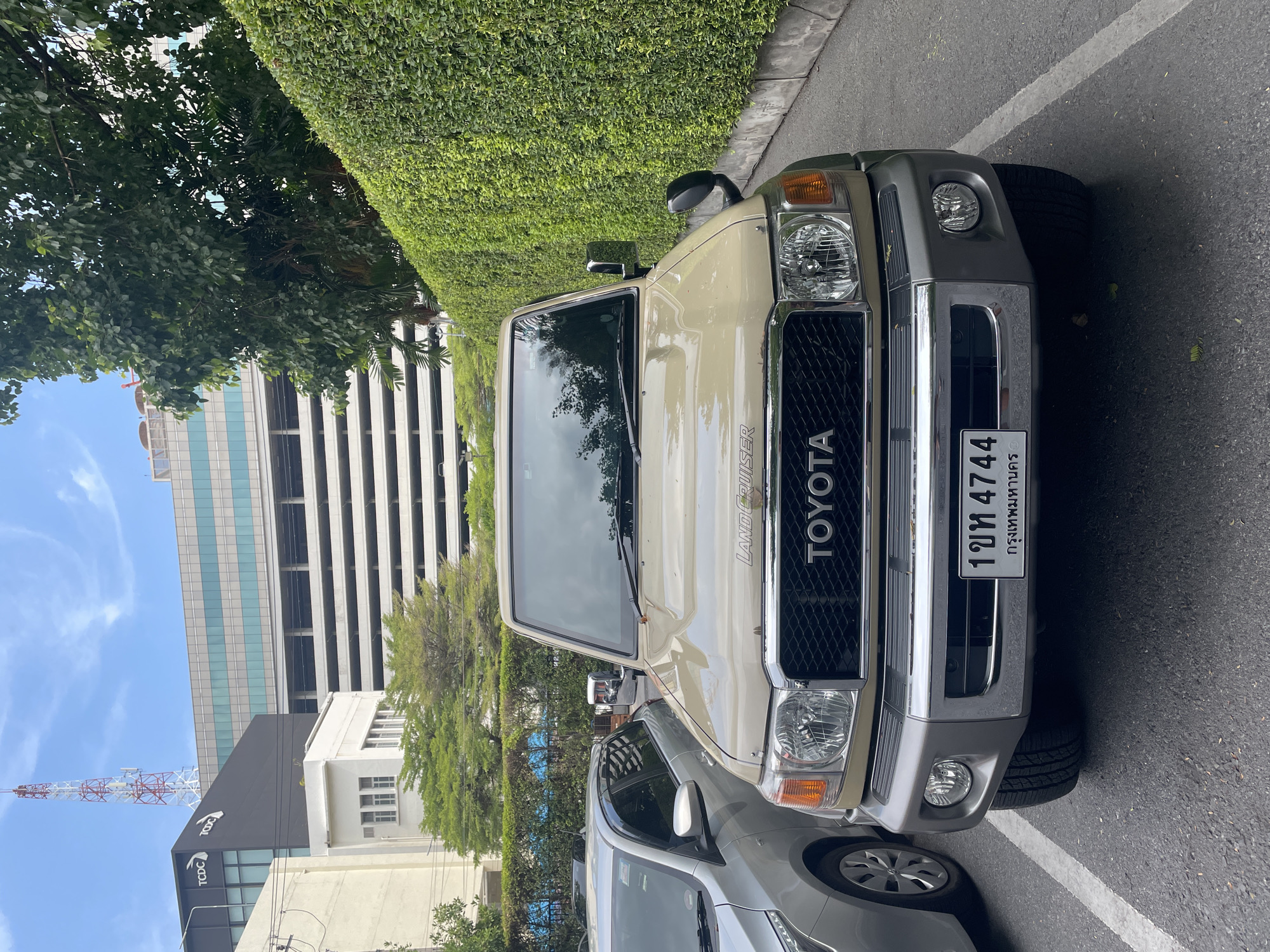 The image shows a beige Toyota Land Cruiser parked on an asphalt surface marked with white parking lines. Behind the Land Cruiser, another car, possibly silver, is partially visible. The Land Cruiser's license plate reads "1ขท 4744 กรุงเทพมหานคร," indicating the car is registered in Bangkok, Thailand. Behind the vehicles is a tall, dense green hedge and a glimpse of an urban landscape, including a multi-story parking garage and a building with the acronym "TCDC" visible, likely the Thailand Creative & Design Center. A communications tower is also seen in the background under a blue sky. The scene is well-lit, suggesting it is daytime. No people are visible in the image.