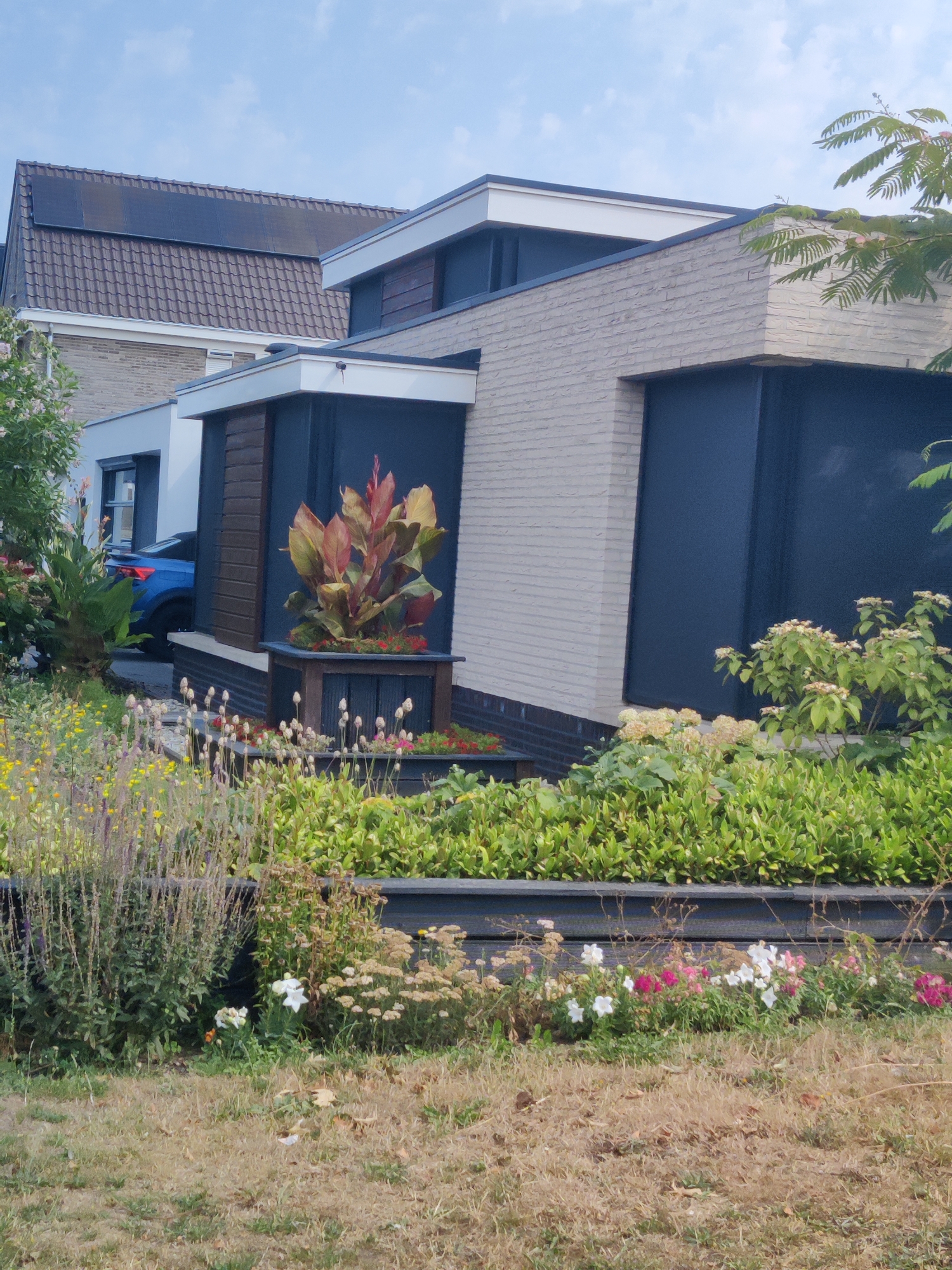 In the city of Winterswijk, Netherlands, a contemporary house with a light brick facade and dark blue accents is visible. Solar panels are installed on the dark tiled roof. In front of the house, a garden features lush green plants and a variety of colorful flowers, including white, pink, and red varieties. A blue car is parked in the driveway. The scene is bathed in natural light under a clear blue sky with scattered clouds.