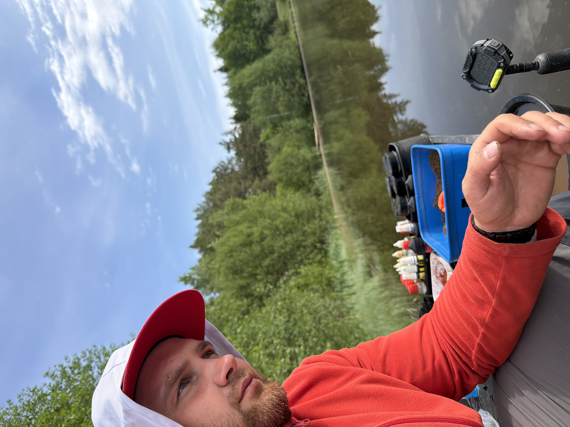 A man with a beard and a red cap is lying down near a body of water. He is wearing a red jacket. A container and some other objects are nearby. Trees and the sky are visible in the background. The location is Prikamskii, Russia.