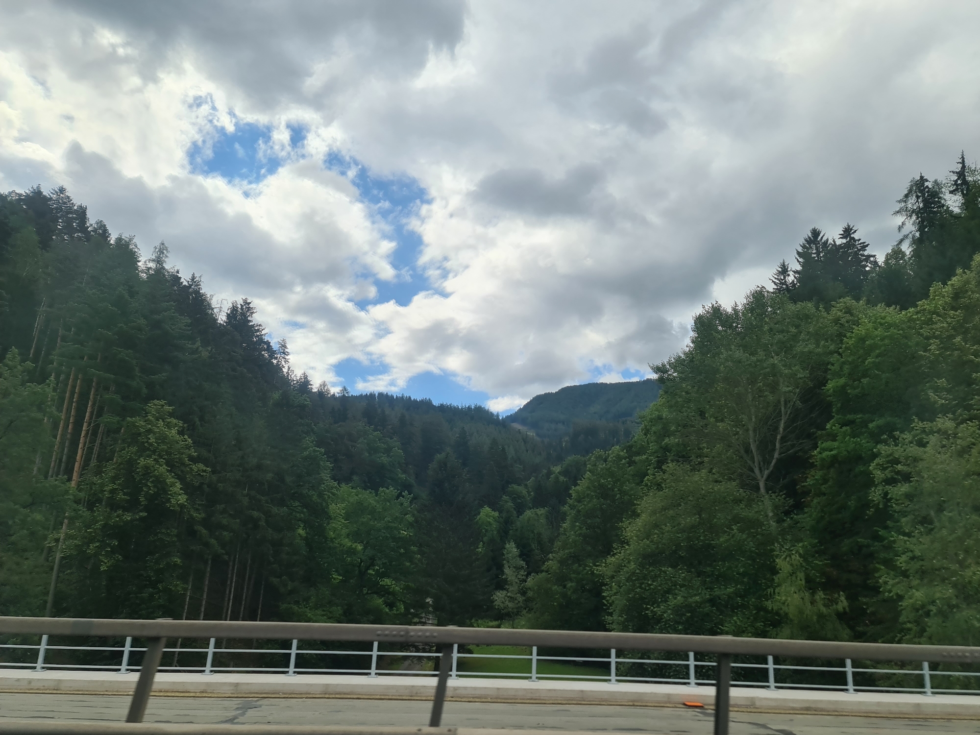 The image depicts a view from a bridge overlooking a lush, tree-covered valley with mountains in the background. The sky is partly cloudy with patches of blue visible. The bridge railing is in the foreground, with a road surface below. This scene is consistent with the landscape found near Leoben, Austria, characterized by its mountainous terrain and dense forests. There are no people or specific actions visible in the image. The environment is natural and serene.