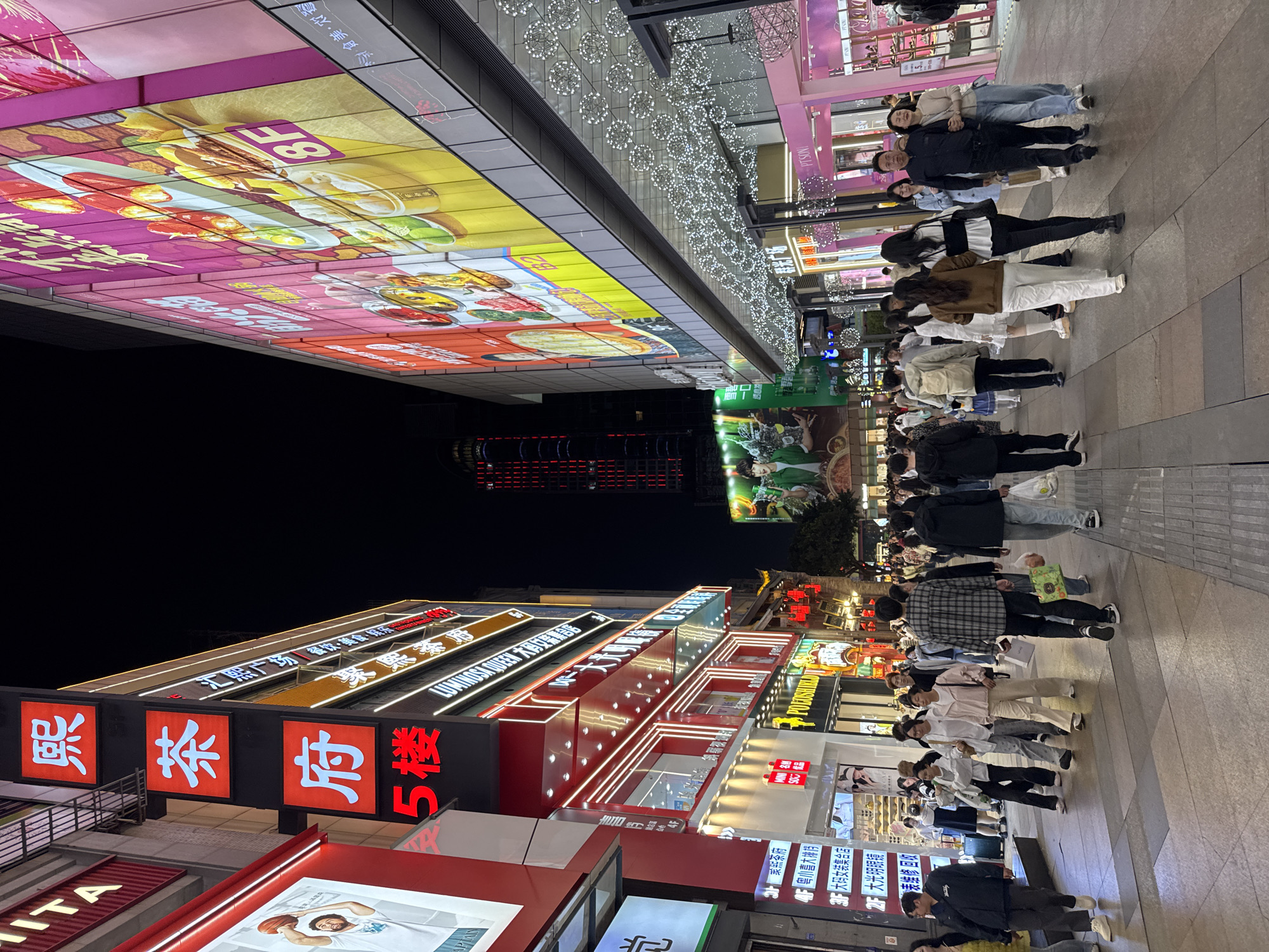 Captured at night in the Jinjiang District of Chengdu, China, the image depicts a bustling commercial street filled with pedestrians. Brightly lit storefronts line the street, with signage in Chinese characters. A group of people are walking along the sidewalk. Overhead, LED displays showcase food advertisements and decorative lighting enhances the ambiance.