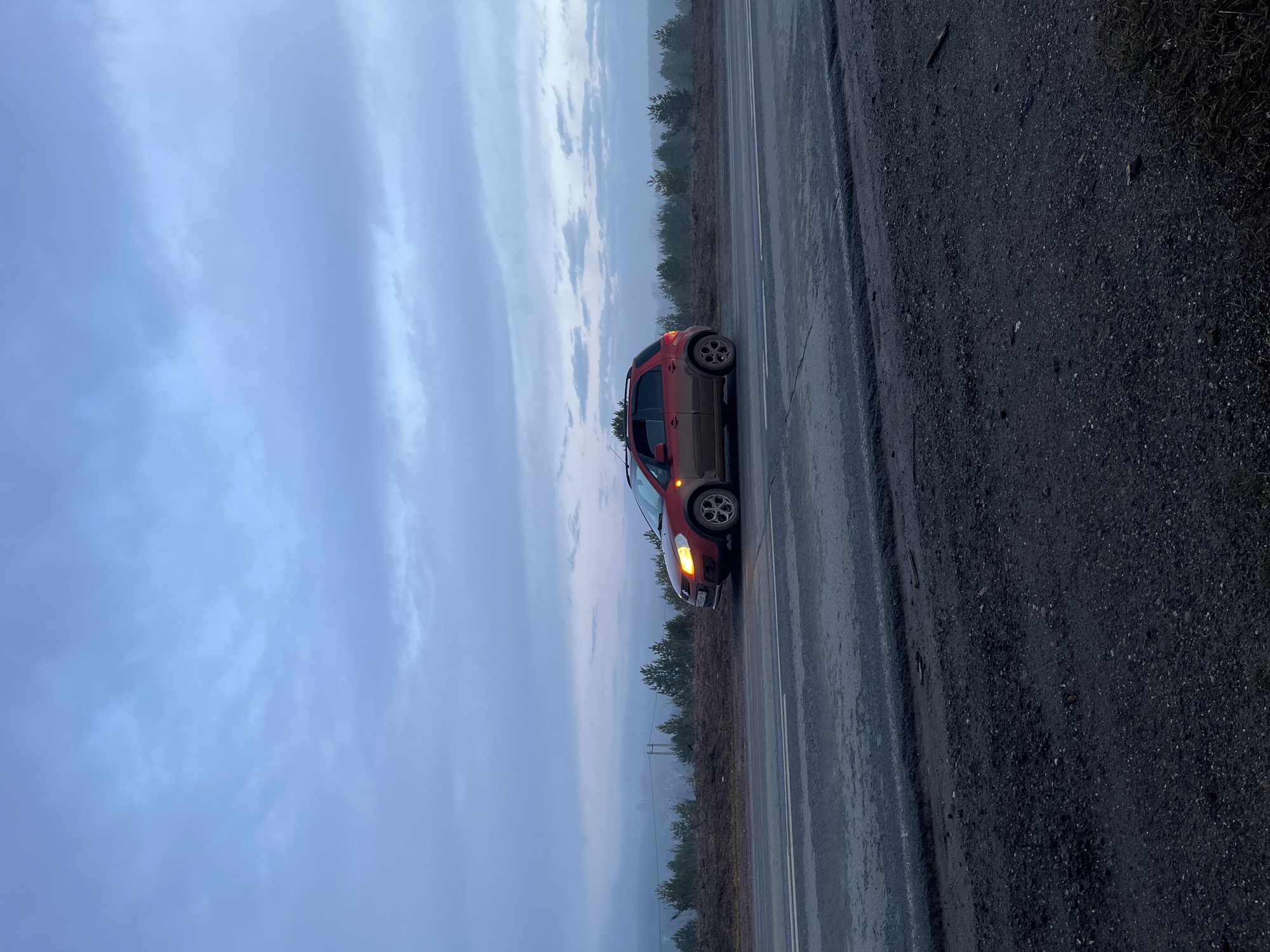 The image shows a red SUV parked on the side of a road in an area with overcast skies and a treeline in the background. The road is wet and appears to be asphalt. The shoulder of the road is gravel. The location is given as Фоки, Russia. The SUV has its front passenger-side indicator light on. There are no people visible in the image.