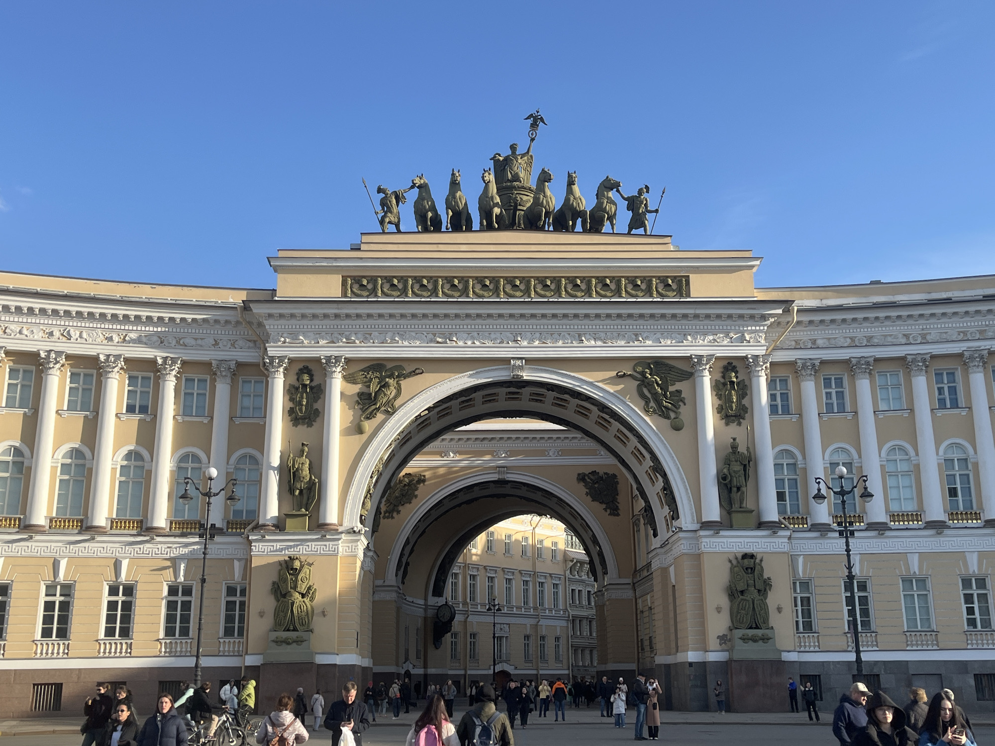 The image displays the Triumphal Arch of the General Staff Building, situated on Palace Square in Saint Petersburg, Russia. The building is a grand Neoclassical structure with a pale yellow facade and white architectural accents, prominently featuring a large central archway.
Atop the arch, a bronze quadriga sculpture is visible, depicting a female figure (Victory) in a chariot pulled by six horses, flanked by two armed figures holding spears. The facade around the arch is adorned with additional bronze statues and relief sculptures, including winged figures. The building features multiple white columns, pilasters, intricate friezes, and both rectangular and arched windows.
The sky overhead is clear and uniformly blue, with no visible clouds. The ground in the foreground is a paved square of a light gray-brown tone. Approximately twenty to thirty pedestrians are scattered across the square and moving through the archway. Individuals are dressed in various forms of outerwear, including jackets and coats, indicating cool weather conditions. Most people are walking, some are observing the surroundings, and a few are engaged with mobile devices. One individual is positioned near a bicycle. A black, multi-lamp street light stands on the right side of the square. Buildings of similar architectural style are visible through the archway in the background. The scene is brightly illuminated by direct sunlight, with some shadows visible on the ground and building details.