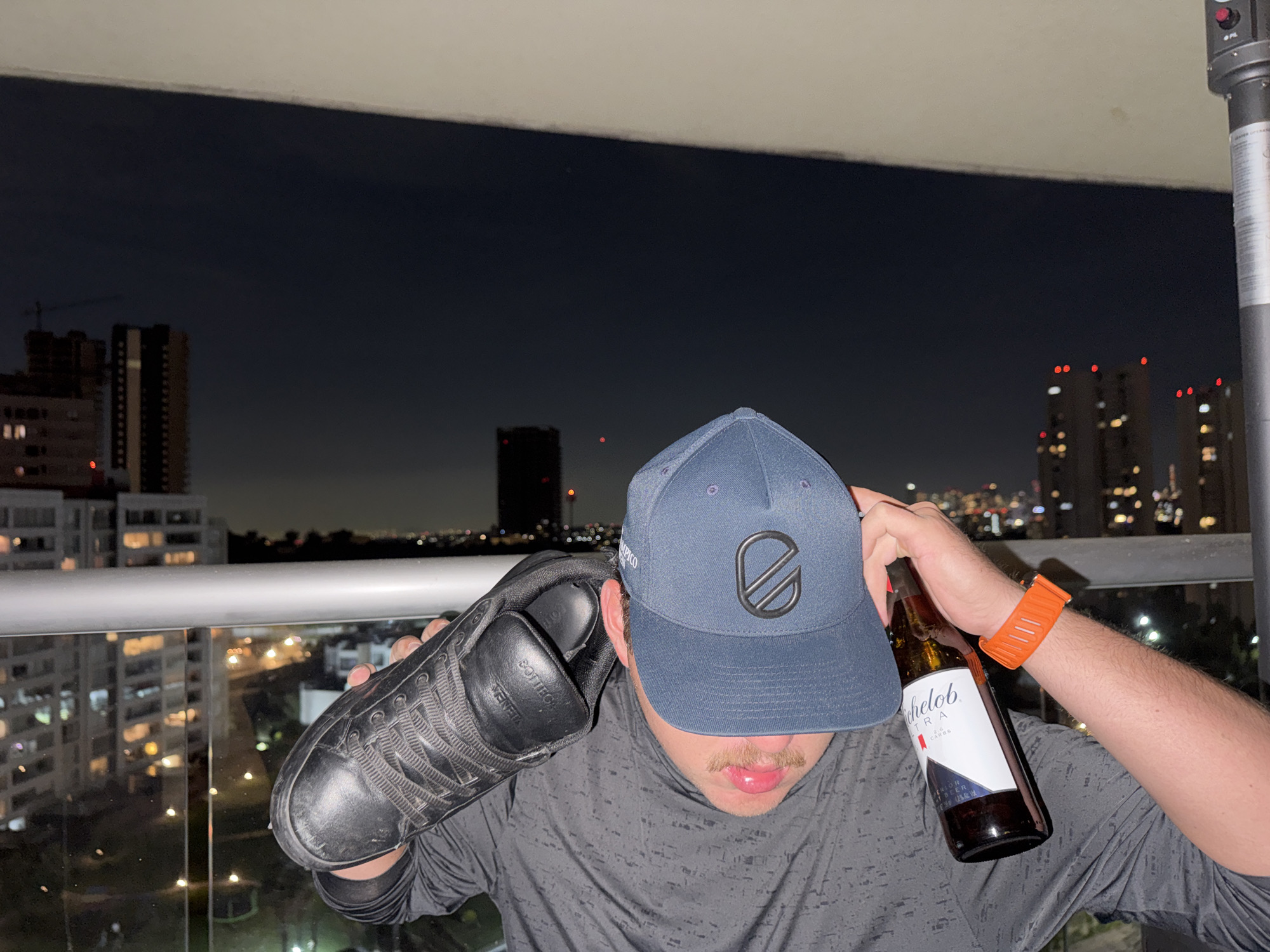A person is on a balcony overlooking the city of Huixquilucan de Degollado, Mexico, at night. The person is holding a black leather sneaker to his ear as if it were a phone and is holding a bottle of Michelob Ultra beer in the other hand. The person is wearing a grey shirt, a blue baseball cap, and an orange wristwatch. In the background, there are buildings and city lights. A metal railing surrounds the balcony.