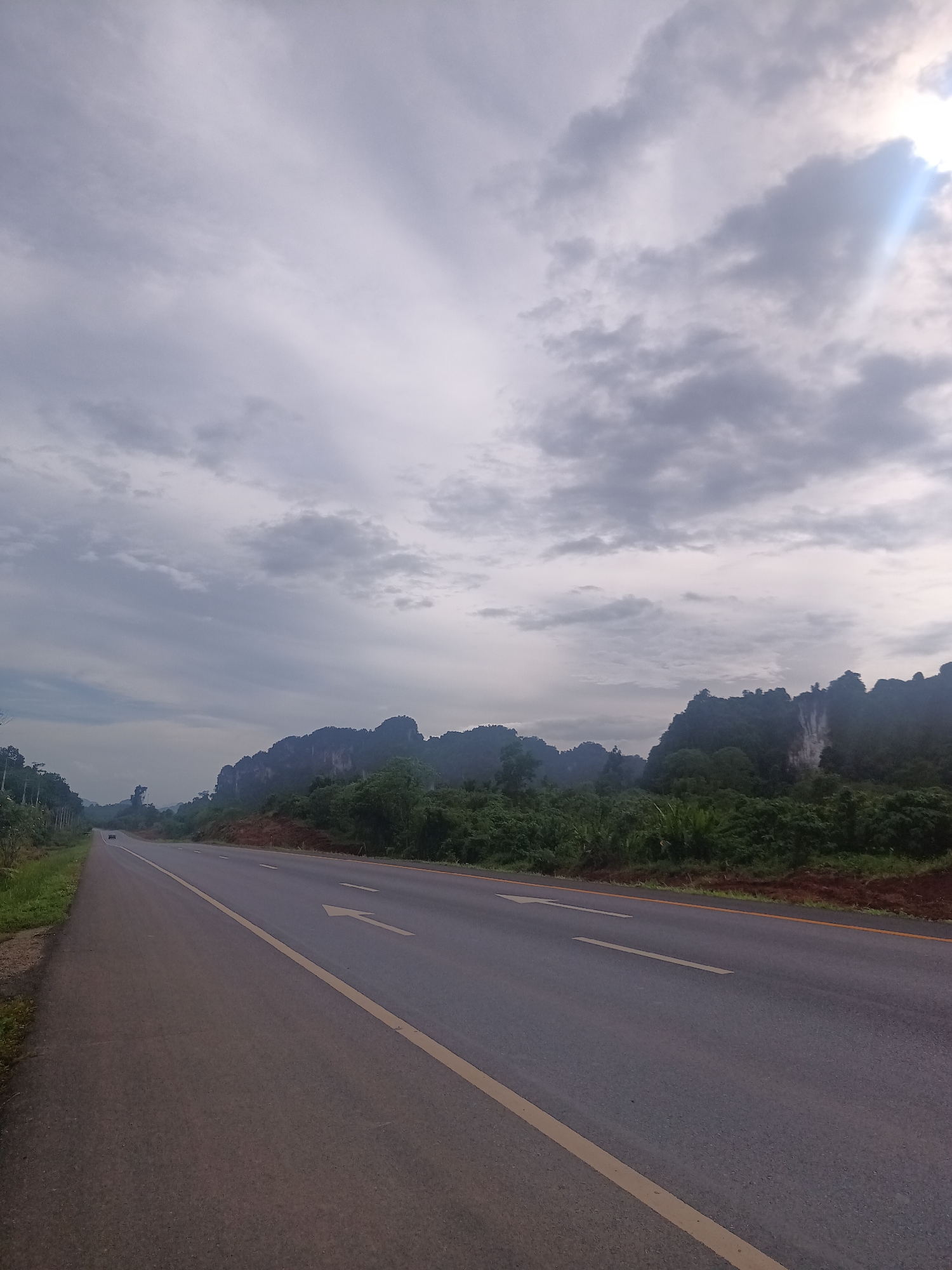 The image shows a road in Plai Phraya, Thailand. The road is asphalt with a yellow center line and a left turn arrow. A vehicle is visible in the distance. Lush green vegetation lines the right side of the road, transitioning to limestone karst formations in the background. The sky is overcast with the sun faintly visible.