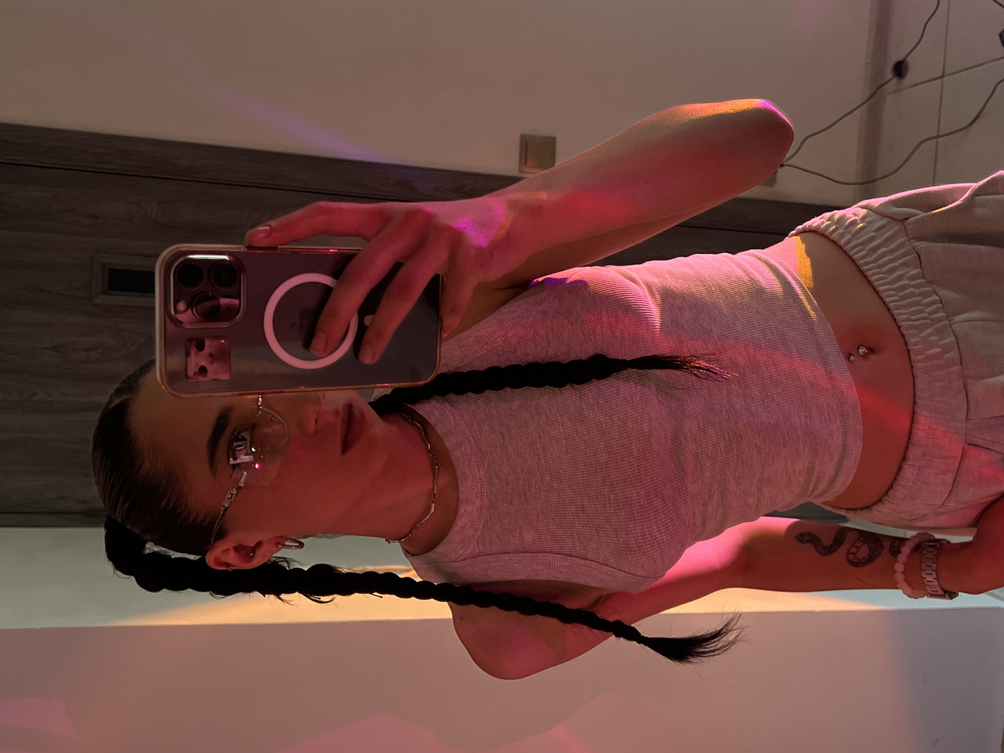 The media file is a selfie taken by a young woman in a room with ambient lighting, casting pink and yellow hues across the scene. The woman is holding a smartphone in her left hand, capturing her reflection. She has dark hair styled in two braids that fall over her shoulders and down her chest. She is wearing a grey ribbed tank top and light-colored sweatpants. She also has on rimless glasses and a silver necklace. A belly button piercing with a silver jewel is visible. On her right arm, there is a snake tattoo and a watch with a silver band and light pink beads. The background appears to be a room with wooden paneling and a white wall. The overall mood is casual and relaxed, possibly taken in the evening due to the lighting. No visible text is present in the image.