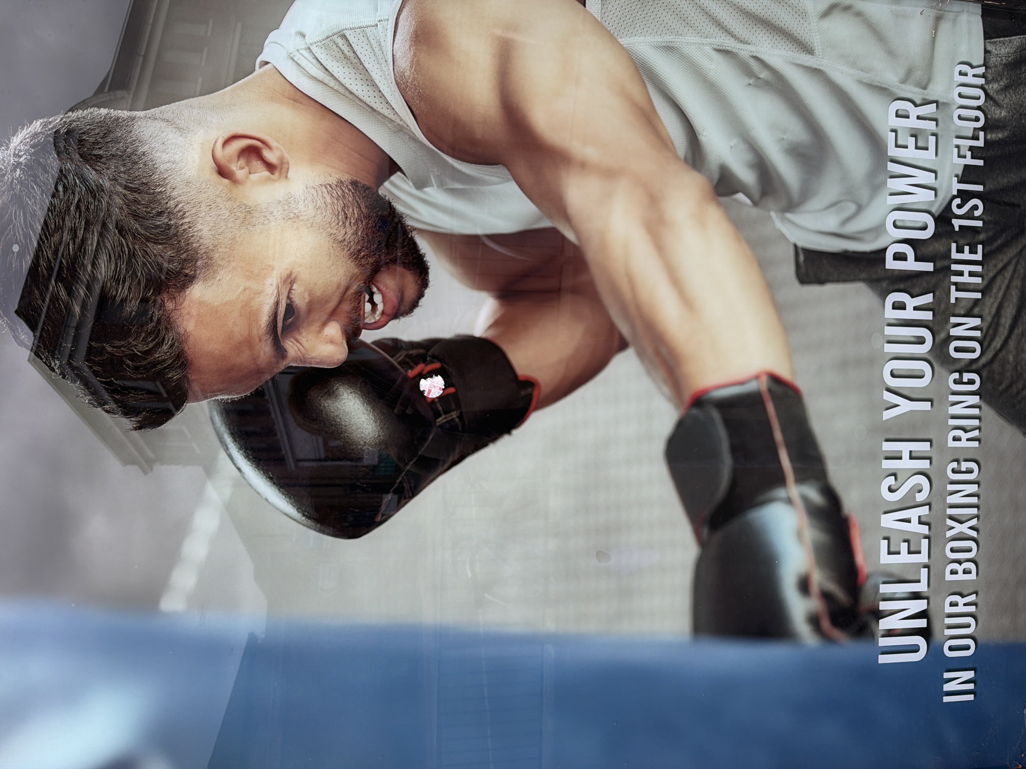 The image shows a promotional advertisement, possibly a poster, featuring a muscular man wearing boxing gloves in a fighting stance. The text on the poster reads "UNLEASH YOUR POWER IN OUR BOXING RING ON THE 1ST FLOOR". The image appears to be taken in a public space, with a blurred reflection of a building visible on the man's head. It is presumed to be in the city of Epsom and Ewell, United Kingdom, based on the instructions.