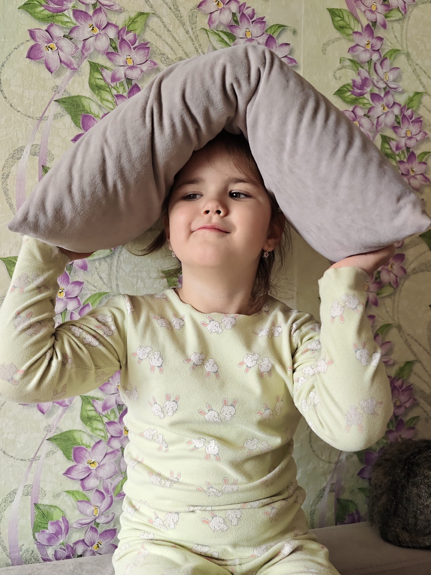 A young girl in a light green pajama set with a white bunny pattern holds a gray pillow over her head indoors, likely in a home in Уральское, Russia. The background features floral wallpaper with purple flowers. A dark-furred animal, possibly a cat, is partially visible in the lower right corner. The girl is looking slightly to her right with a small smile. She wears star-shaped earrings. The overall lighting suggests an indoor setting.