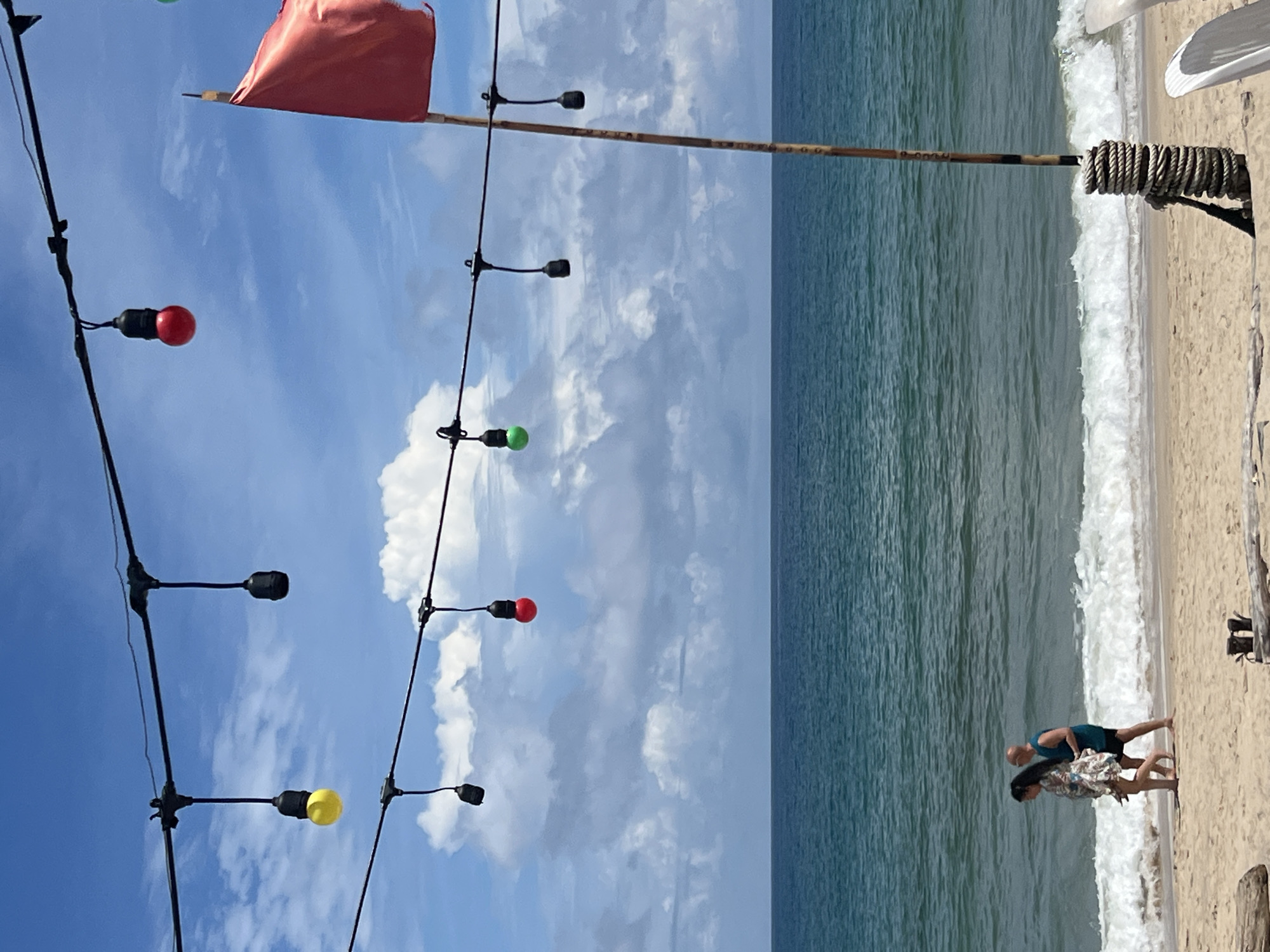 In Choeng Thale, Thailand, on a sandy beach, a couple walks along the shoreline. The ocean, with a moderate tide, stretches to the horizon under a bright, blue sky dotted with fluffy white clouds. Overhead, a string of decorative lights hangs, with several bulbs lit in yellow, red, and green. A red flag flutters on a wooden pole on the right side of the frame, secured with coiled rope. The beach is strewn with driftwood in the foreground, and the scene evokes a relaxed, tropical atmosphere.