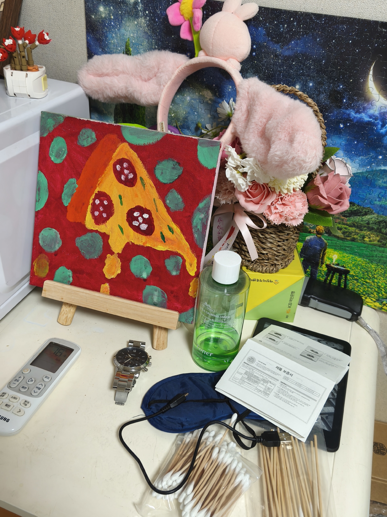 The image depicts a cluttered tabletop scene, likely in a residential setting in Asan-si, South Korea, as suggested by the presence of Korean text on various items.
In the foreground, a white Samsung air conditioner remote control displays a temperature of "18". To its right, a silver-banded wristwatch with a black face rests on the table. Adjacent to the watch is a small, dark blue pouch with a black USB cable attached, and a clear plastic bag filled with wooden cotton swabs.
Dominating the mid-ground is a vibrant, amateur painting of a slice of pizza with red and green dots on a red background, displayed on a small wooden easel. To the right of the painting, a green plastic bottle of "Calming Toner" stands next to a woven basket overflowing with artificial flowers in shades of pink and white. Behind the flowers, a yellow box with Korean writing is partially visible.
In the background, a large, colorful jigsaw puzzle depicting a night sky with a crescent moon and a pastoral landscape is leaning against a wall. To the left of the puzzle, a white pot with red artificial flowers is positioned on a white surface, possibly a shelf or dresser. Further left, a pink fuzzy headband with bunny ears is visible.
The overall impression is one of a personal space filled with decorative items, personal care products, and electronics. The lighting appears to be indoor artificial light. No individuals are present in the image.