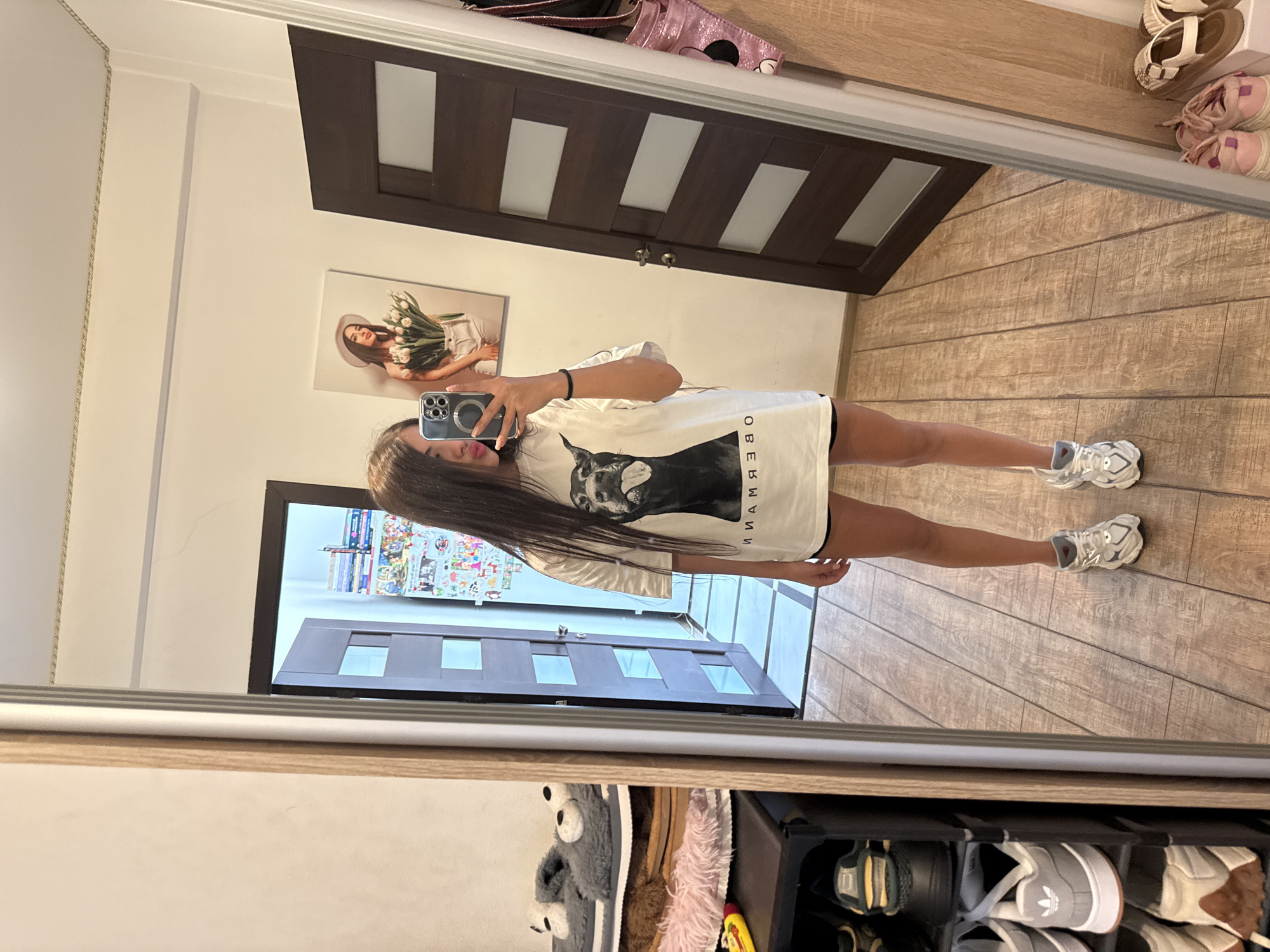 A young woman stands in a hallway, taking a mirror selfie. She has long, dark hair and is wearing a white t-shirt with a black dog graphic and black shorts. She has on white and grey sneakers. To her left, a shoe rack holds several pairs of shoes and slippers. To her right, a dark wood door with glass panels is visible. In the background, a poster of a woman holding tulips hangs on the wall. The flooring is wood-look tile.