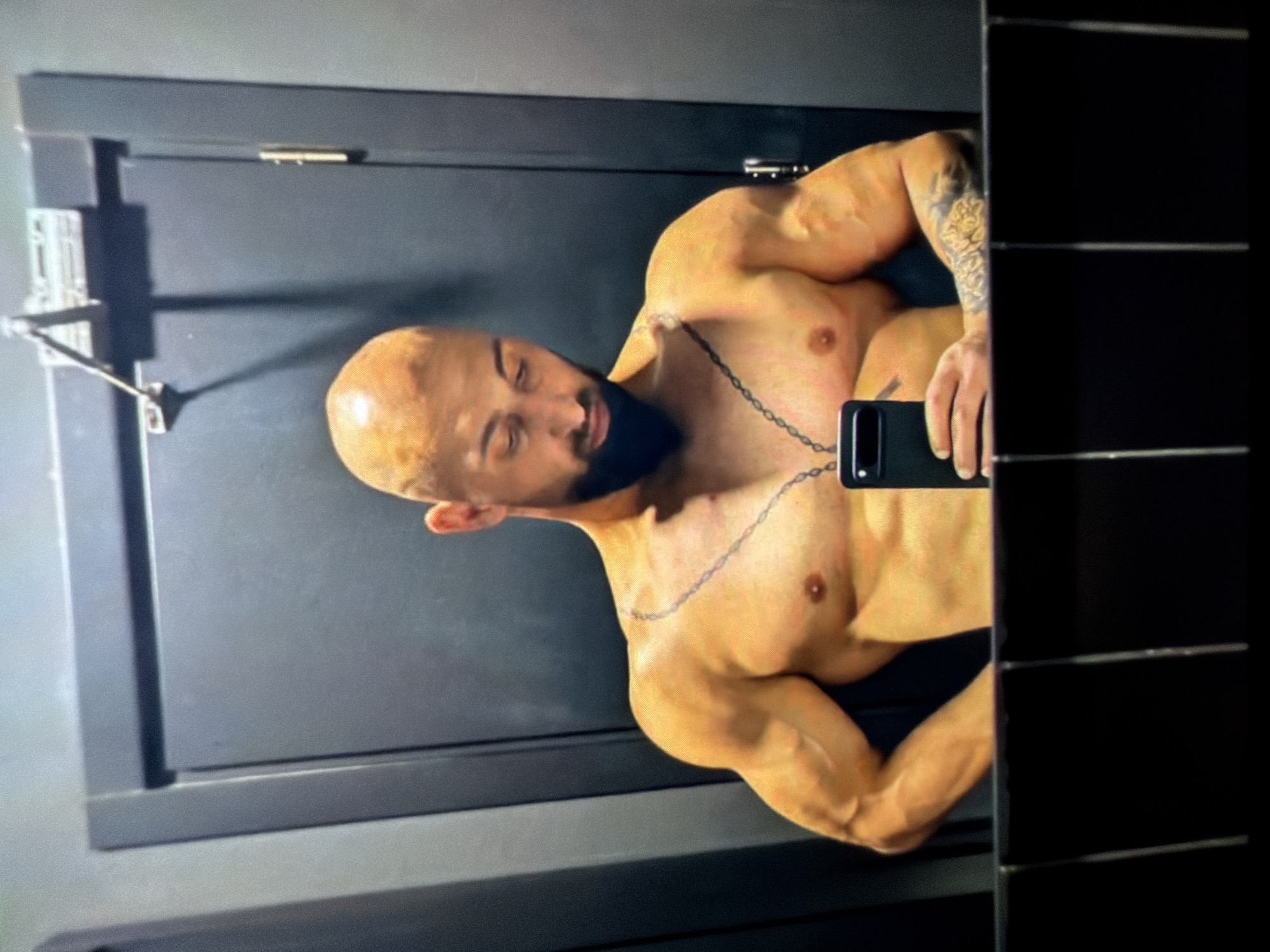 A shirtless, muscular man with a bald head and a beard is holding a black smartphone, presumably taking a selfie in a mirror. He has a chain necklace and tattoos on his right arm. The reflection shows he is standing in what appears to be a bathroom with a dark-colored door or wall behind him and horizontal tiles on the right side. The exact location in the city of Unknown, Unknown is not identifiable from the image.