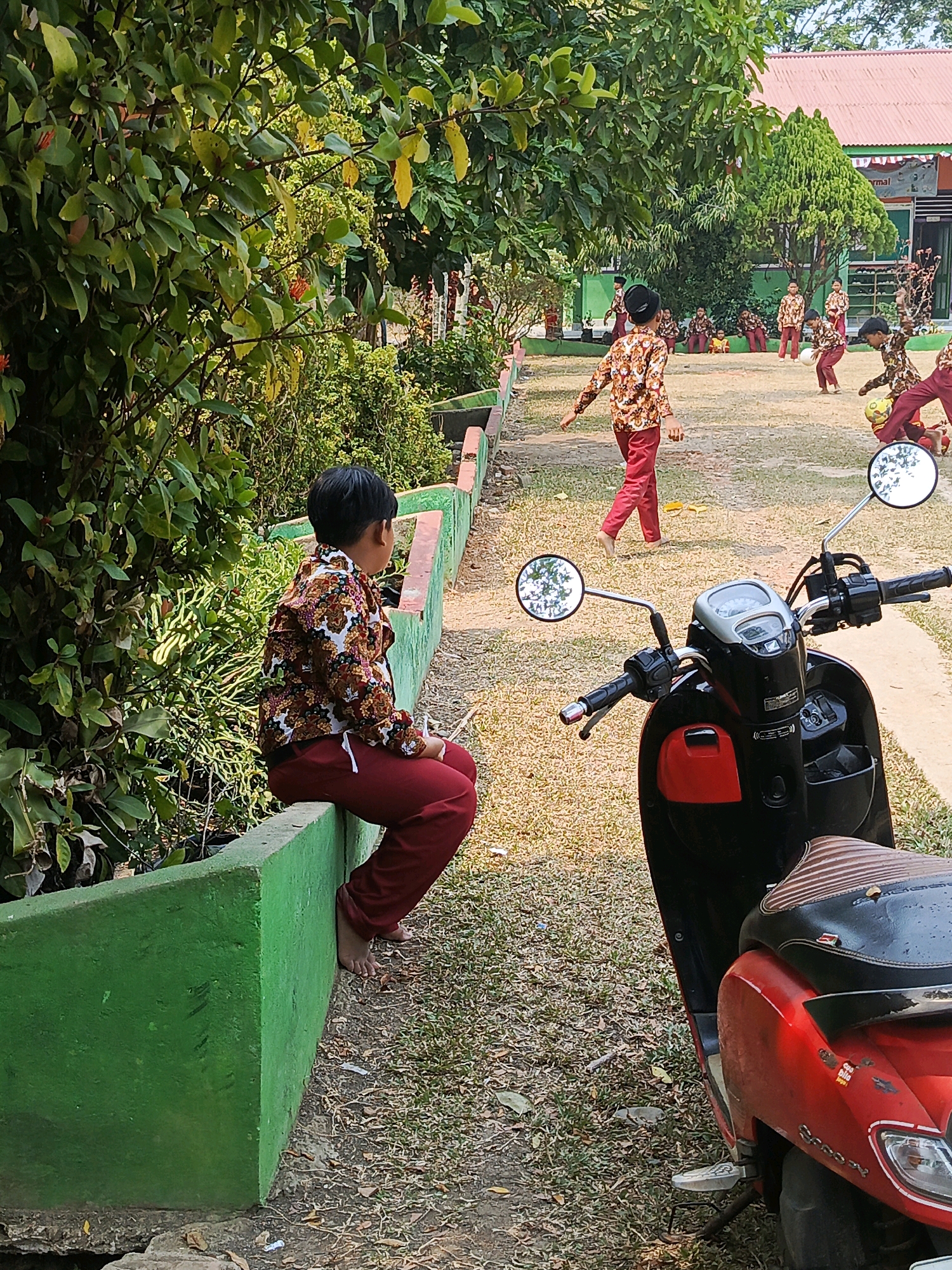 In Megang Sakti IV, Indonesia, a young boy wearing a patterned shirt and maroon pants sits barefoot on a bright green concrete planter box, facing away from the camera. He holds a red object in his right hand. In the background, other children in similar attire are playing soccer on a grassy field in front of a building with a red roof. A red and black scooter is parked in the foreground, to the right of the boy. The environment is outdoors, with trees and foliage surrounding the playing area. The sun is shining, casting shadows and illuminating the scene.
