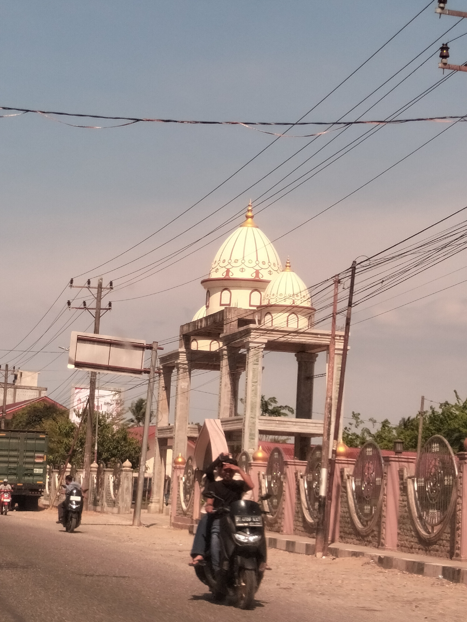 The scene captures a street in Meudangara, Indonesia. In the foreground, a person on a black scooter passes by on the street. Further down the road, another person rides a scooter behind a green truck. The architecture is visible as a decorated fence and a building with two white domes, supported by columns. Overhead, numerous power lines intersect the sky. The sky is a muted blue, suggesting either early morning or late afternoon light.