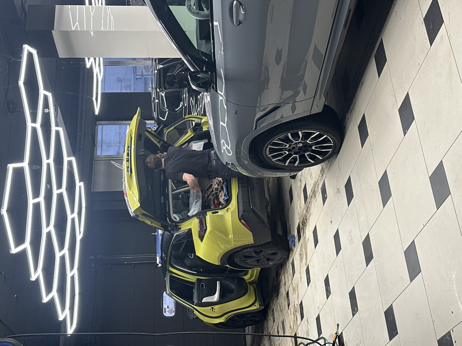 The scene is set inside an auto detailing facility, identified as АвтоД-110, located at Obukhovskoy Oborony Ave, 110, Building 2, Saint Petersburg, Russia. The ceiling is dark, illuminated by a network of hexagonal LED light fixtures arranged in a honeycomb pattern. The floor is tiled with a checkerboard design of light and dark squares, showing signs of moisture and tire tracks.
Two SUVs are prominent. A bright lime-green SUV, with its rear hatch and driver's side rear door open, occupies the left-center of the frame. A male individual, seen from behind, with short dark hair, wearing a black short-sleeved shirt and dark pants, is positioned at the open rear of the lime-green SUV, appearing to access or arrange items within the trunk, including a visible leopard-print fabric object and a white, wrapped package. To the right, a grey SUV, likely a BMW M model identified by its distinctive wheel design and a red 'M' emblem on the fender, is partially visible. A darker SUV is discernible in the background, behind the grey vehicle. A black hose or cable lies on the floor on the far left. A window in the background reveals an overcast outdoor environment.