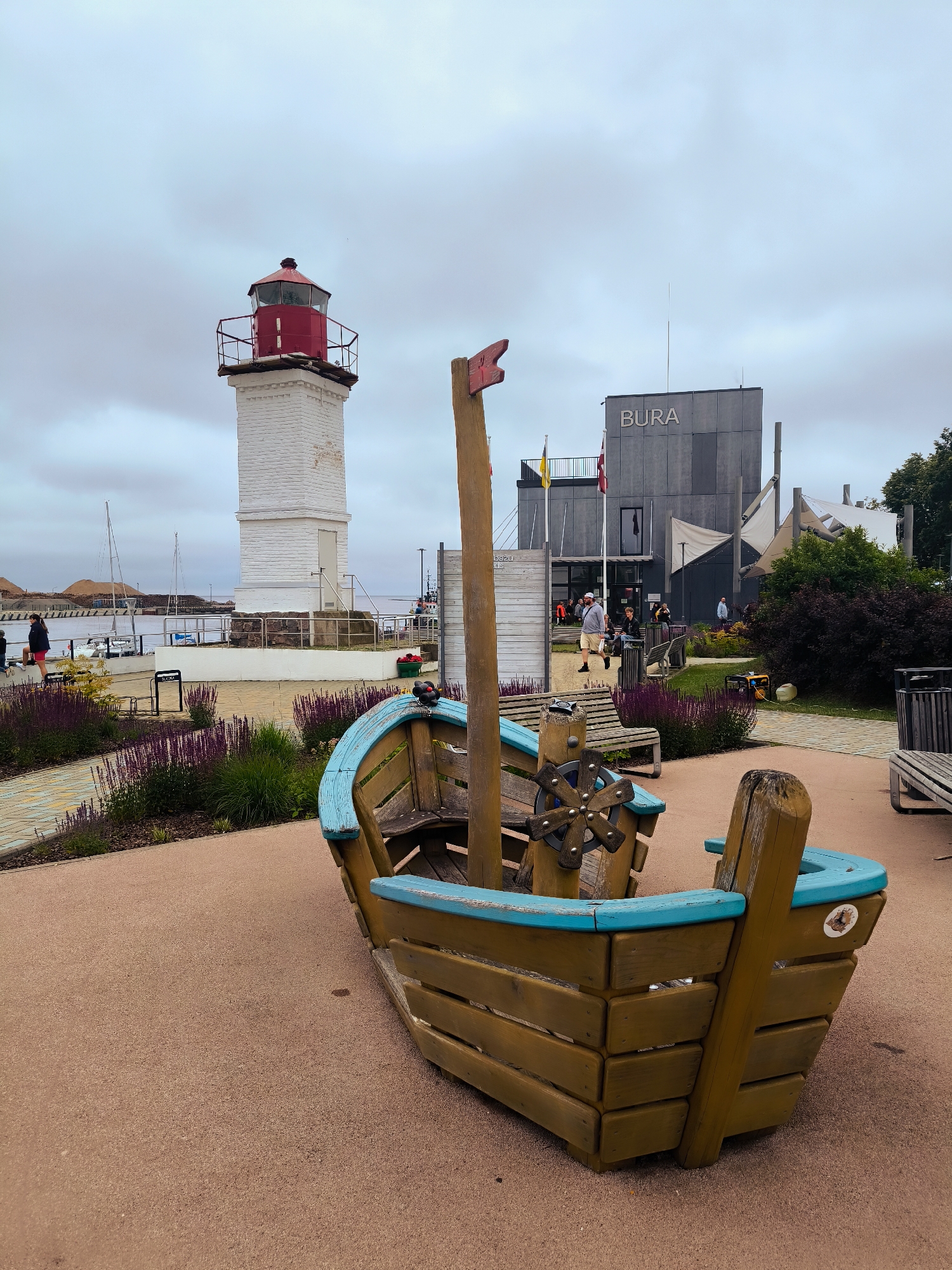 In Salacgrīva, Latvia, a wooden boat-shaped play structure sits on a sandy ground in the foreground. Behind it, a pathway lined with purple flowers leads towards a white lighthouse with a red top. Further back, a modern grey building with the word "BURA" inscribed on it stands prominently, adorned with flags and awnings. Several people are visible walking around the building and on the pathway. Sailboats are docked in the harbor to the left of the lighthouse. The sky is overcast, suggesting a cloudy day.