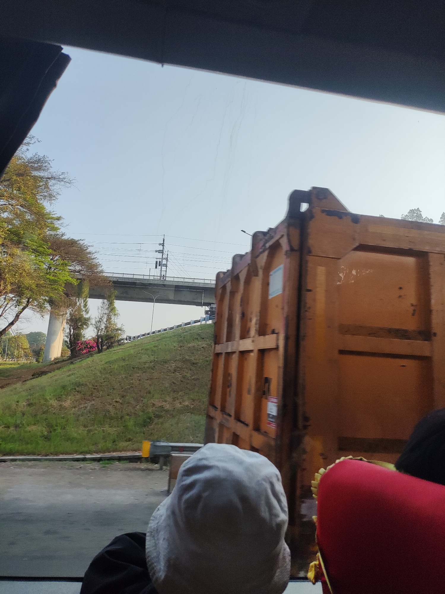 The image shows a view from inside a vehicle looking out. In the foreground, the back of a person wearing a light-colored head covering and a dark shirt is visible on the left. To the right, a red seat is partially visible. Outside the vehicle, a large, orange dumpster is prominently featured, partially obstructing the view of the surrounding environment. Beyond the dumpster, a grassy slope leads up to an elevated road or bridge. Trees with green and brown foliage are scattered along the slope and to the left of the bridge. The sky is clear and pale blue, with some faint lines or streaks visible. Utility poles and wires are present above the bridge. A yellow marker post is visible on the road behind the dumpster.