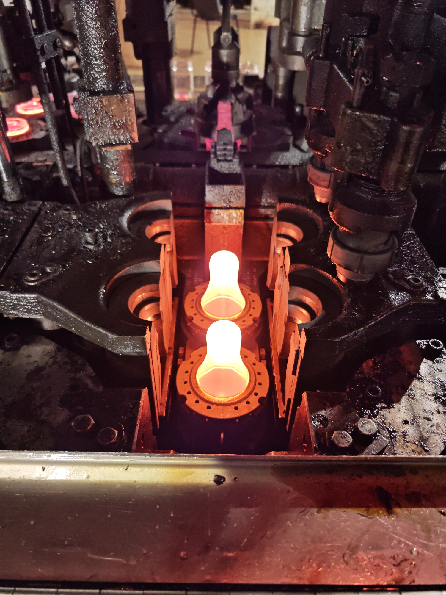 The image depicts a section of an industrial glass-blowing machine, likely within a factory setting in Pleven, Bulgaria. Two glowing, bulbous glass shapes are positioned within metallic molds or supports. The machine components are heavily lubricated, with a dark, oily residue coating the metal surfaces. Mechanical arms or actuators are visible above and to the sides of the molds. The environment is dimly lit, with the primary source of illumination emanating from the molten glass itself. There are no people visible in the frame. The machine appears to be in operation, heating and shaping glass.