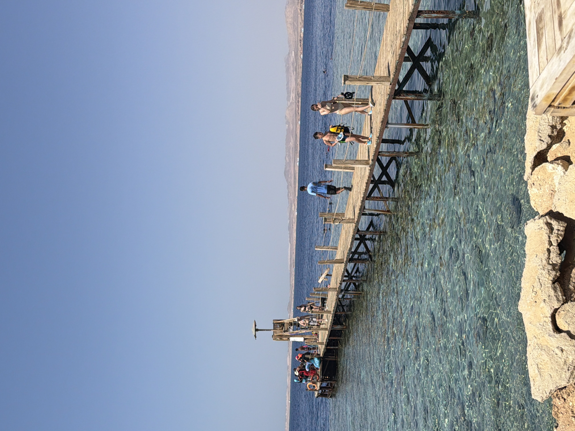 The image depicts a wooden pier extending into the clear blue waters of the Red Sea. The pier is a popular spot for tourists in Sharm El Sheikh, Egypt, as evidenced by the presence of several individuals engaged in various activities. On the left side of the pier, a group of people are gathered near a small platform, some are sitting, others standing. Further along the pier, a few people are walking, some in swimwear, suggesting they are either heading to or from the water for swimming or snorkeling. In the background, a sandy coastline with arid hills is visible, characteristic of the Sinai Peninsula. The water is exceptionally clear, revealing the coral formations beneath the surface, a common sight in the diving and snorkeling destinations around Sharm El Sheikh. The sky is a clear, bright blue, indicating a sunny day.