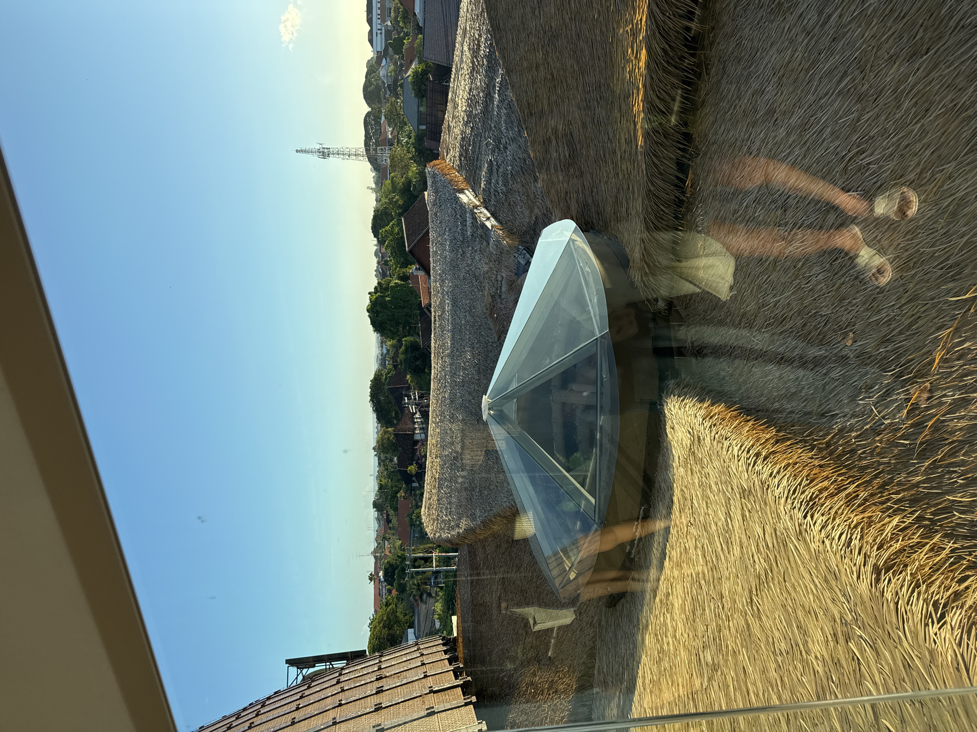 The image displays an outdoor scene from a high vantage point, likely a window or balcony, overlooking a series of rooftops under a bright, clear sky. The photo is rotated 90 degrees clockwise, but described as if oriented correctly.
In the immediate foreground, a large, reflective glass surface dominates the lower right, showcasing the reflection of a person's lower body, specifically their legs and feet in sandals. This suggests an individual standing near the window, looking out. The glass also reflects some of the textured roofs below.
The mid-ground is characterized by an array of rooftops. Many are traditional thatched roofs, showing layers of dried straw or palm fronds, indicative of tropical architecture. Sunlight warmly illuminates these roofs, highlighting their texture and creating subtle shadows. On one of these thatched structures, a contemporary, multi-faceted glass skylight or dome-like feature is visible. Further in the scene, some buildings have more conventional tiled roofs, with a section of solar panels visible on one such roof in the lower left.
Beyond the buildings, a landscape of dense green trees extends into the distance. A tall metal lattice tower, possibly a communications or transmission tower, stands prominently against the horizon. The sky is a gradient of clear light blue, transitioning to a yellowish hue near the horizon, indicative of either early morning or late afternoon lighting conditions. The weather appears sunny with minimal cloud cover. The architectural styles and lush greenery align with the context of a tropical location, such as Denpasar, Indonesia. No discernible text is visible in the image.
