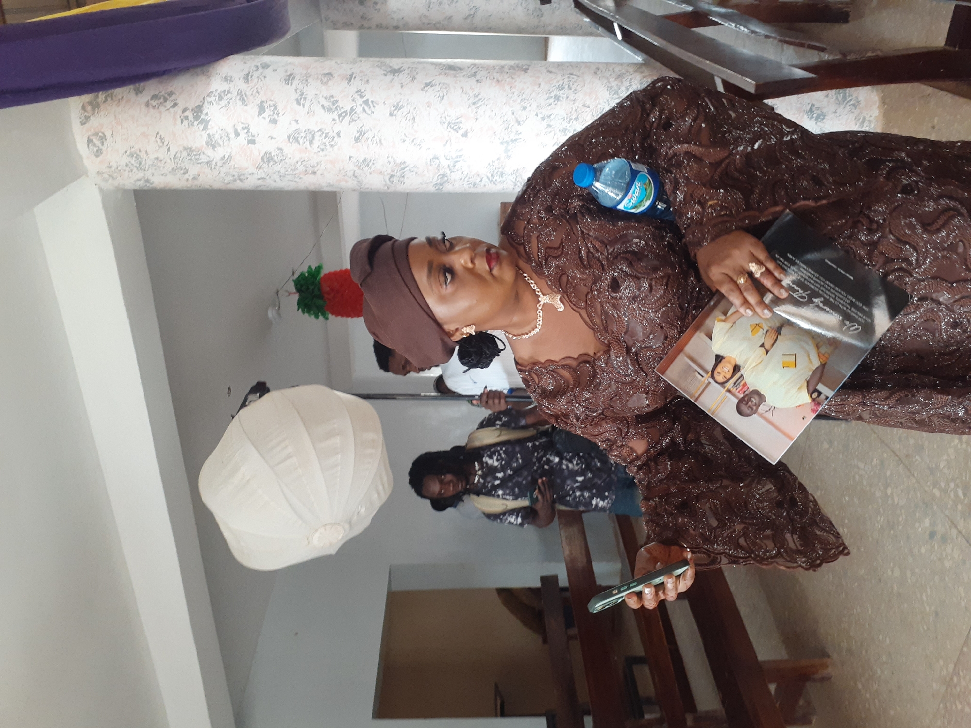 A medium-shot depicts a woman in a brown dress holding a photo book. The woman is wearing a brown head wrap, gold necklace, and earrings. She is holding a cell phone in one hand. She has a water bottle on her right side. In the background, there are other people and a white paper lantern hanging from the ceiling. The location is in a city called Unknown, Unknown.