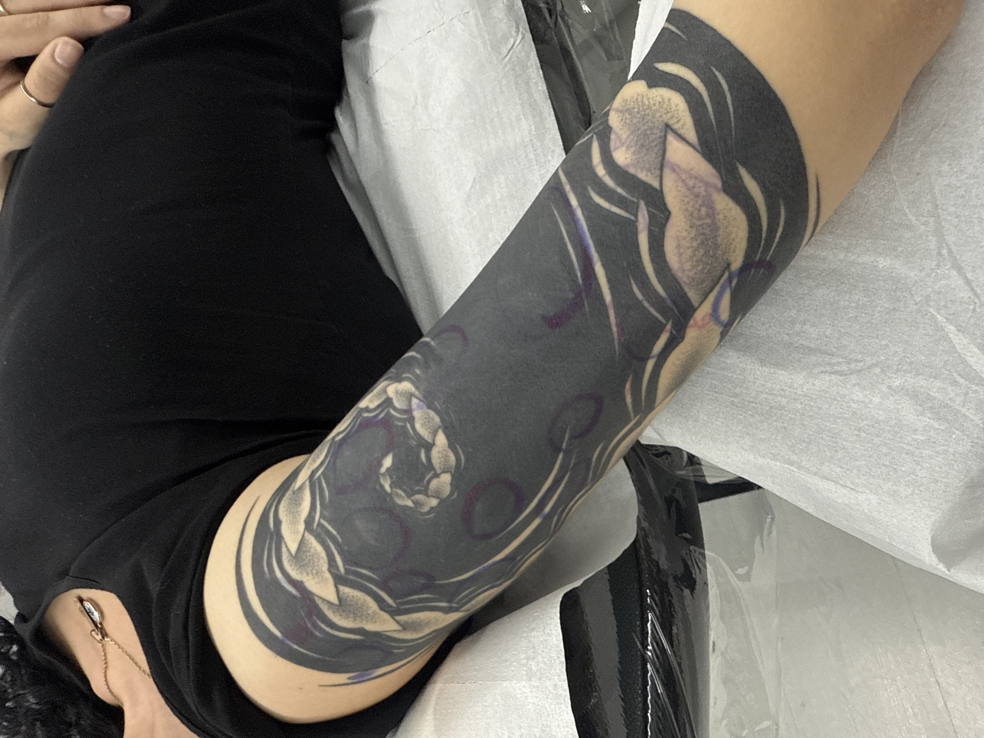 The image shows a close-up of a person's arm with a large, dark tattoo sleeve. The tattoo features intricate designs with lighter elements against a dark background, possibly depicting organic or floral motifs. The person is wearing a black top and has jewelry, including a ring and a necklace. They are lying on a white, possibly paper-covered surface, which could be a medical or tattoo artist's table. There is no information in the image that indicates the city or location within Sofia, Bulgaria.