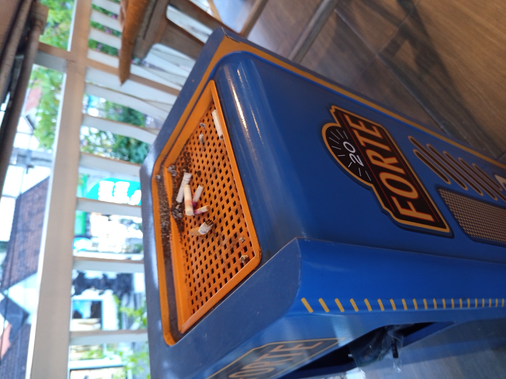 A close-up shot of a blue cigarette disposal container with an orange grate top filled with cigarette butts. The container is labeled "FORTE 20" in bold, dark brown lettering. The container's blue exterior has yellow markings. The environment suggests an outdoor setting in North Kuta, Indonesia, with a partial view of a stairway or building structure with plants visible in the background. The focus is on the container and its contents.