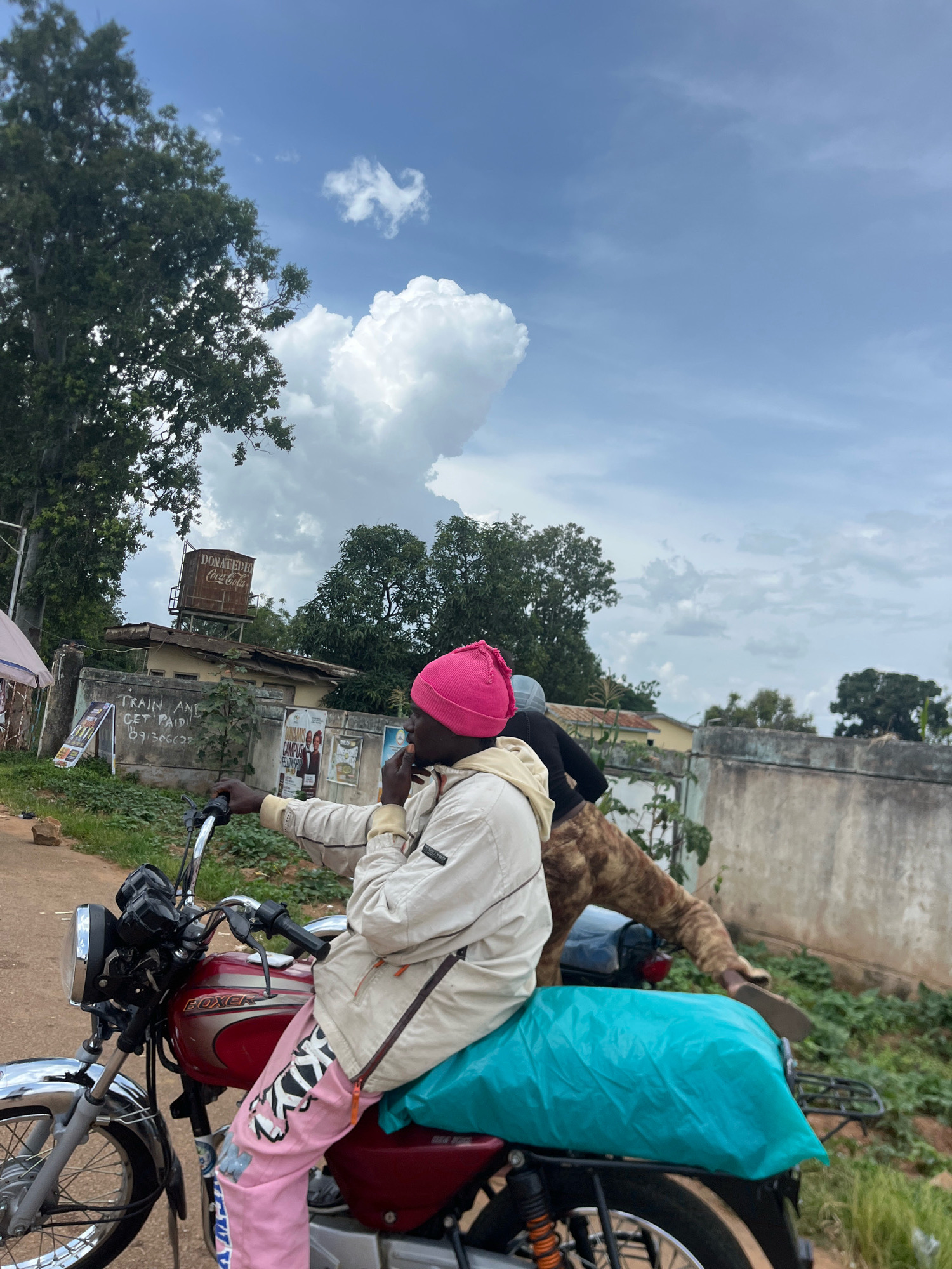 Two individuals are positioned on a red motorcycle in a roadside environment. The person in the foreground, wearing a pink beanie and a light-colored jacket, is seated on the motorcycle and appears to be holding something to their mouth. They are also wearing pink pants with black and white lettering. Behind them, another individual is also seated on the motorcycle, wearing a patterned jacket. A turquoise-colored bag is secured to the back of the motorcycle. The background reveals a concrete wall with posters, trees, and a water tank with the inscription "DONATED BY Coca-Cola." This scene is located in the city of Rafin Par, Nigeria. The sky above is partly cloudy.