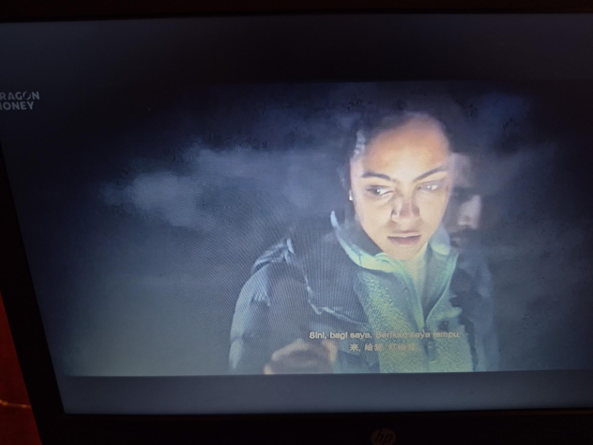 The image shows a screen displaying a scene with two people. The person in the foreground is a woman with dark hair, looking intensely towards the left. She is wearing a teal jacket or hoodie. In the background, a man's face is partially visible, appearing to be looking in the same direction as the woman. The overall scene is dark and hazy, with some atmospheric effects suggesting smoke or mist. White text overlays the image at the bottom, with some of the text appearing to be in a non-English script. In the top left corner of the screen, there is text that reads "DRAGON MONEY". The screen itself is a display, likely a television or computer monitor, with a dark bezel and an HP logo visible at the bottom edge. There is no indication of the location being in the city of Sandwell, United Kingdom.
