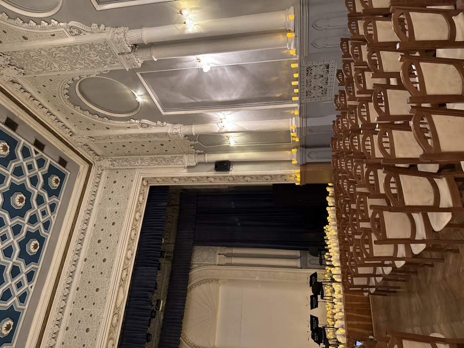 The image displays the Grand Hall of the House of Scientists (Kyiv House of Scientists of the National Academy of Sciences of Ukraine), located at Volodymyrska Street 45A in Kyiv, Ukraine. The hall is set up for a performance, characterized by an empty audience area and an illuminated stage.
In the foreground and mid-ground, numerous rows of uniform wooden chairs with light-colored upholstered seats and backs are arranged on a polished wooden parquet floor, oriented towards a stage. No individuals are present.
The stage, situated on the left, features a dark blue main curtain. To the left of the stage opening, a large white screen or backdrop is visible. Several black music stands, some marked with "BLACK TIE" branding, are positioned on the stage, along with a few black chairs. The stage floor and a tiered platform directly in front of it are densely covered with hundreds of glowing yellow-orange electric candles. Additional electric candles line the lower wall ledges along the right side of the hall.
The hall's interior architecture exhibits a Moorish Revival style. Walls are adorned with intricate white plasterwork featuring geometric and floral motifs, pilasters, and arched recesses. Multiple wall-mounted light fixtures with glass shades illuminate the space. The ceiling is decorated with a distinct pattern of dark blue and lighter blue geometric shapes, incorporating star-like elements and gold accents. The ambient lighting is warm and subdued, primarily emanating from the electric candles and wall sconces.