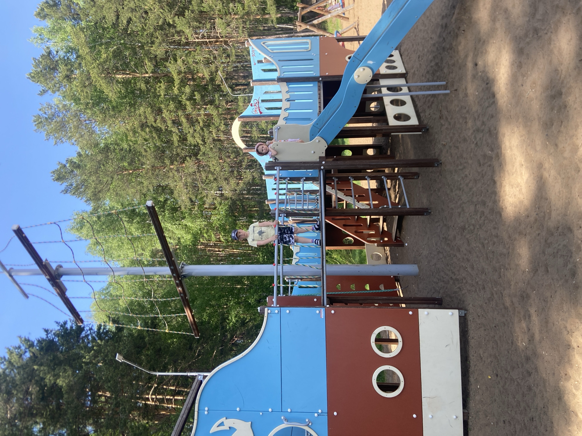 A playground in Chaikovsky, Russia, features a large, colorful structure resembling a pirate ship with blue and brown panels, a mast with netting, and a slide. Two children are present: one stands on the ship's deck wearing a light-colored shirt, dark shorts, and a baseball cap; the other sits at the top of the slide wearing a pink hat. The playground surface appears to be sand. Evergreen trees surround the play area, and a blue sky is visible.