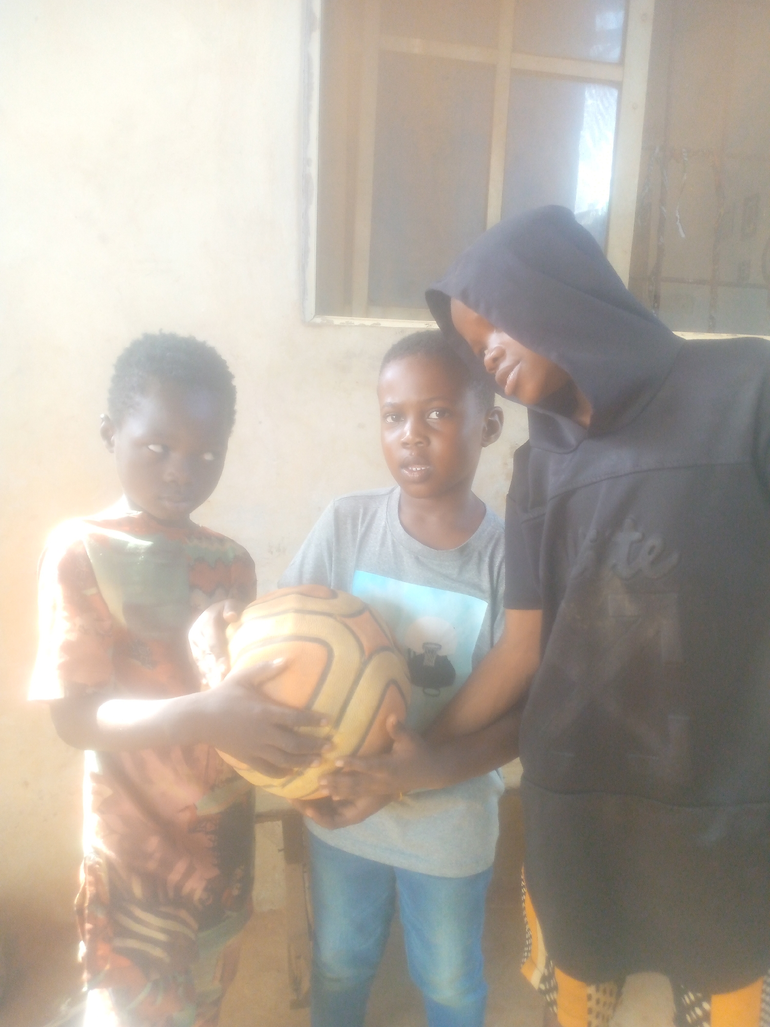 Three children stand outdoors, holding a basketball. The child on the right wears a dark-colored hooded sweatshirt and patterned yellow and black pants. The child in the center wears a grey T-shirt and blue jeans. The child on the left wears a patterned shirt. A window is visible in the background. The location is Unknown, Unknown.