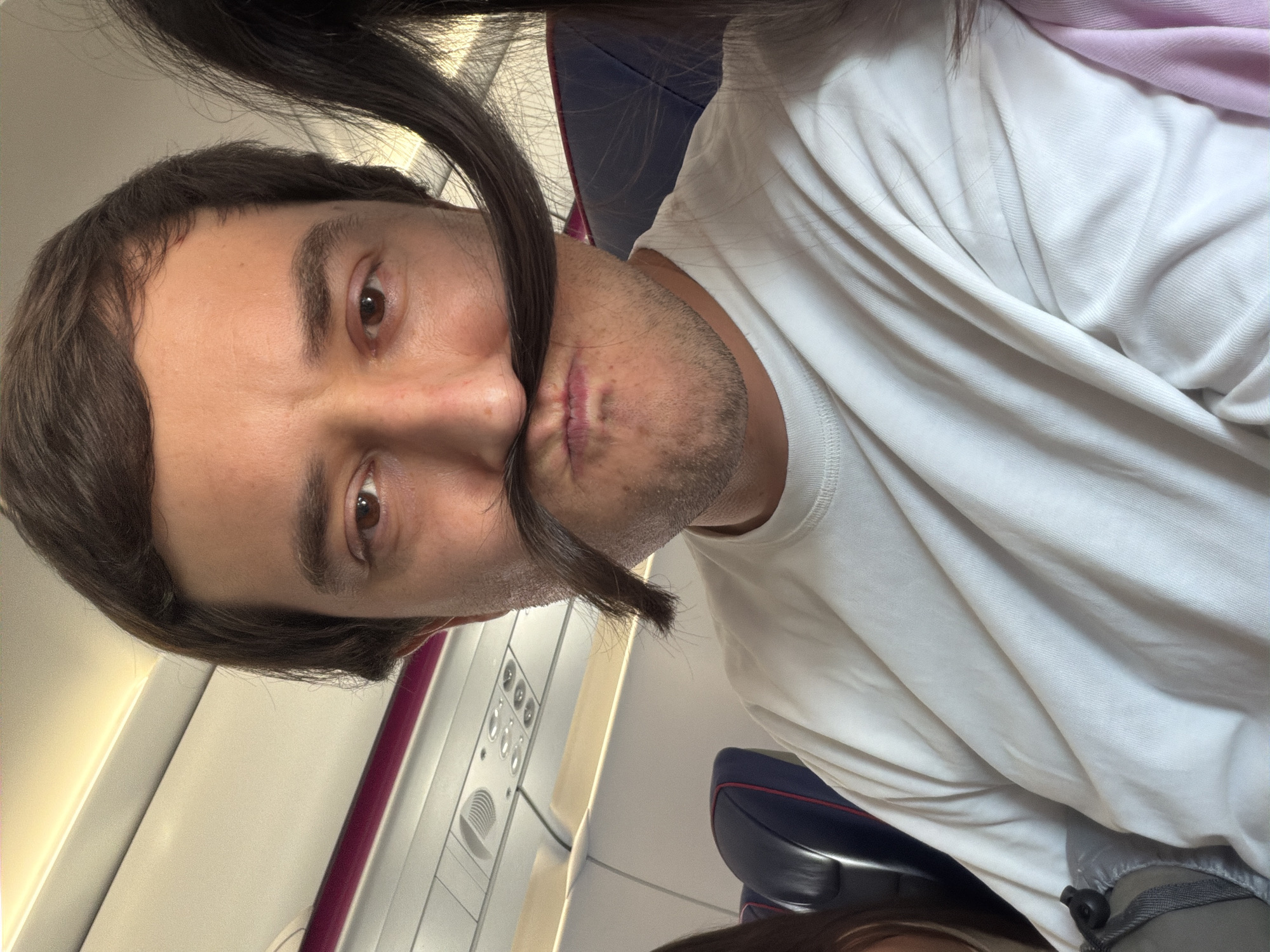 The image shows a close-up, selfie-style portrait of a man sitting on an airplane. He has brown hair and is wearing a white t-shirt. His mouth is pursed and he has a strand of dark hair across his upper lip, creating the appearance of a mustache. In the background, airplane seats and overhead compartments are visible. The scene is lit by artificial cabin lighting and natural light from a window. The man appears to be in motion, as indicated by the slight blur of his hair and the angle of the photograph. There is no indication of the city of Donja Cijevna, Montenegro in this image.