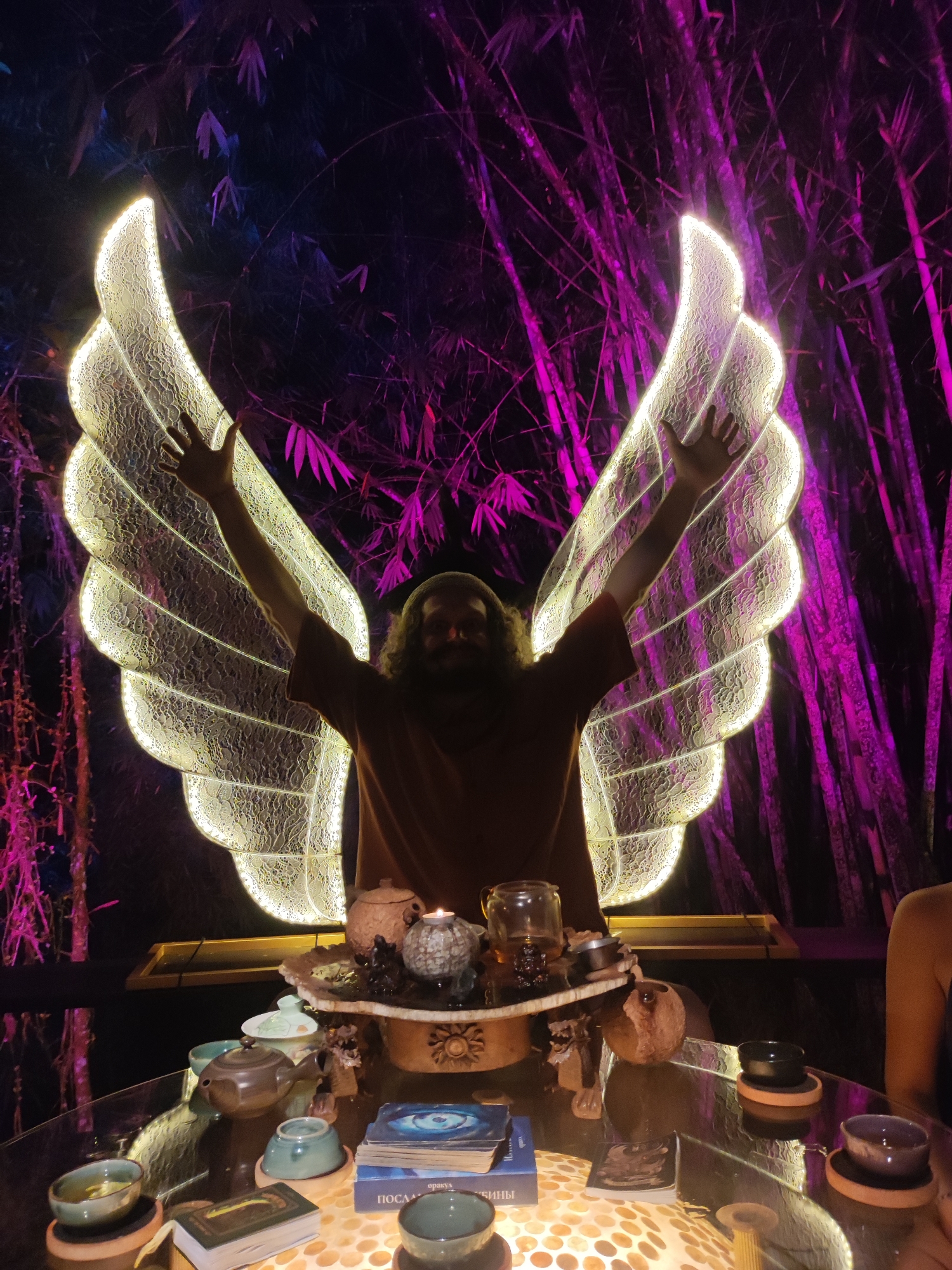 A man with long hair and a beard stands with his arms raised in front of illuminated wings. He is wearing a brown t-shirt. A table in front of him holds teacups, teapots, tarot cards, a candle, and other decorative items. The table is on a surface that is illuminated from below. Behind the man and wings is a backdrop of bamboo stalks lit with purple light. The scene suggests an outdoor setting at night, possibly in a decorative garden or tea ceremony area in Lokaserena, Indonesia.