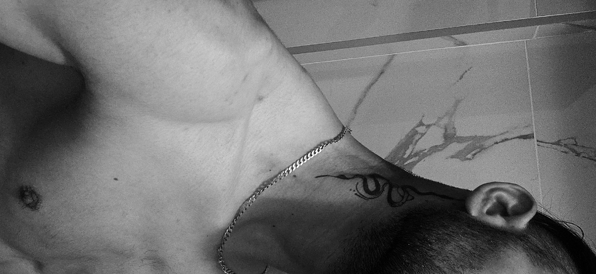 A monochrome, low-angle shot shows a person's upper chest, shoulder, neck, and head in what appears to be a bathroom setting. The person has a visible pectoral muscle and a light gold chain resting across their collarbone. A tattoo of a stylized figure is visible on the right shoulder. The ear is prominent on the right side of the frame, with short dark hair visible above it. The background consists of what appears to be bathroom tiles with a marble-like pattern. There are no other people or objects present in the frame. The location in Plovdiv, Bulgaria is undetermined from this image.