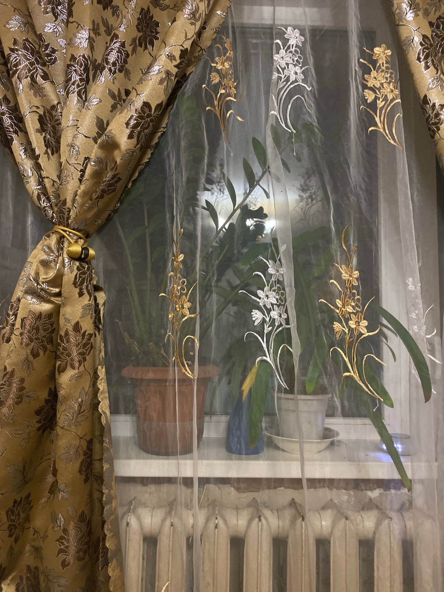 The image shows a window with curtains and plants. A thick, golden curtain with a floral pattern is draped to the left, held back by a gold tie. A sheer white curtain with embroidered floral designs in gold and white hangs in front of the window. Several potted plants are on the windowsill. A beige radiator is visible below the windowsill. The scene is likely indoors, possibly in a residential setting, in Коробицыно, Russia. There are no people or animals visible.
