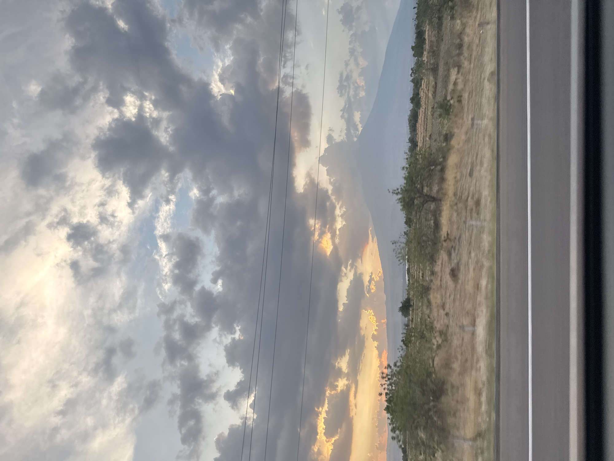 The image depicts a landscape scene taken from inside a vehicle, possibly a car or bus. The perspective is through a window, with part of the vehicle's interior visible on the right side. Outside the window, there is a view of a field or open area with dry grass and some vegetation. Beyond that, a mountain range is visible in the distance. The sky is filled with clouds, some of which are dark and appear to be associated with a sunset or sunrise, as they have a golden glow around the edges. Power lines stretch across the sky.
It's difficult to pinpoint the exact location in Metepec, Mexico without additional context or reference points in the image. The description focuses on the visible elements within the frame.