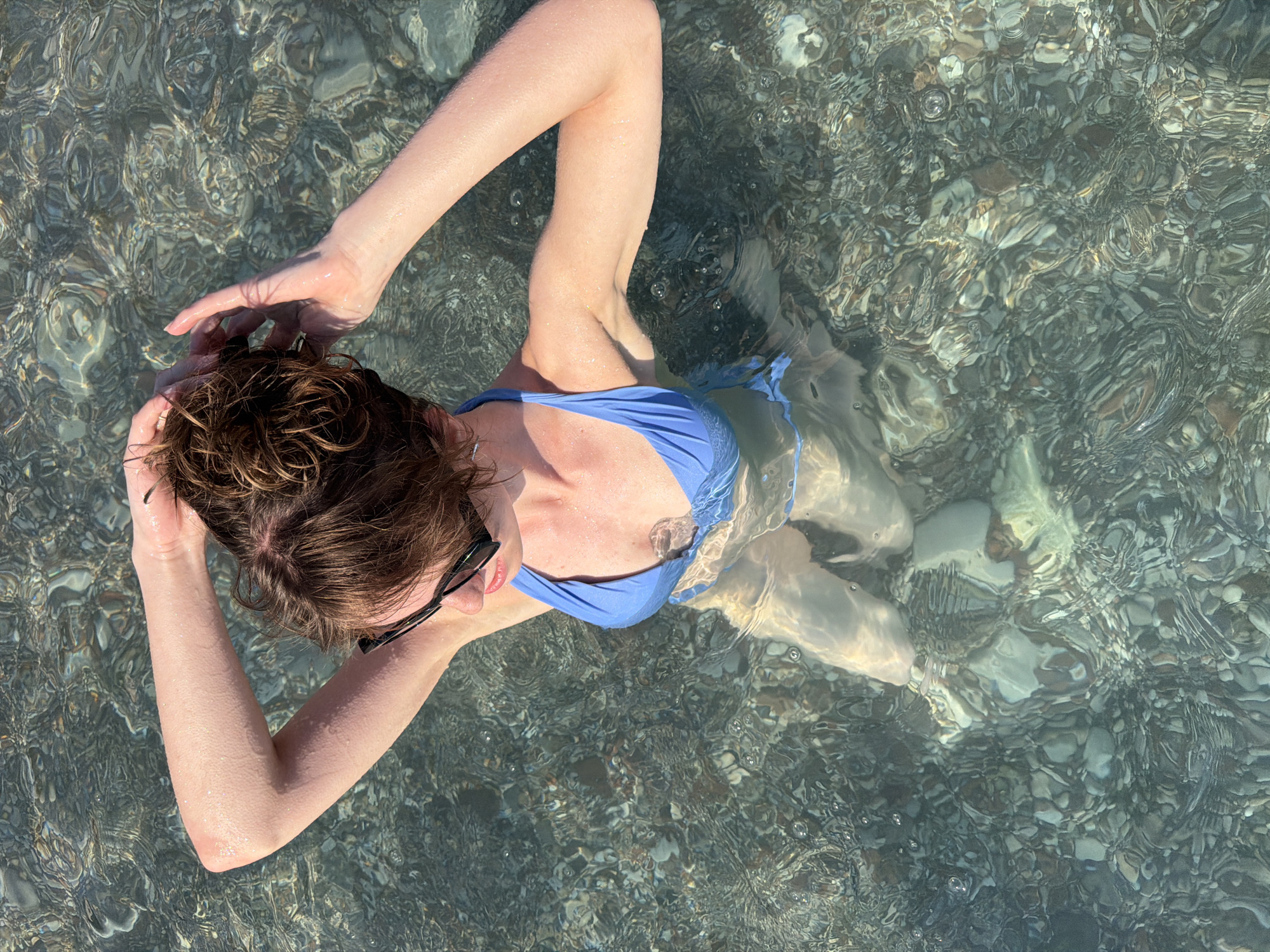 A high-angle, medium shot shows a young woman with brown hair pulled up in a bun, wearing black sunglasses and a blue bikini, standing in clear, shallow water at the beach in Sutomore, Montenegro. She is looking down and touching her hair with both hands. The seabed is visible beneath the water, covered with smooth, multi-colored pebbles. Sunlight reflects on the water's surface, creating distorted patterns. The water is calm, with a few small bubbles visible.