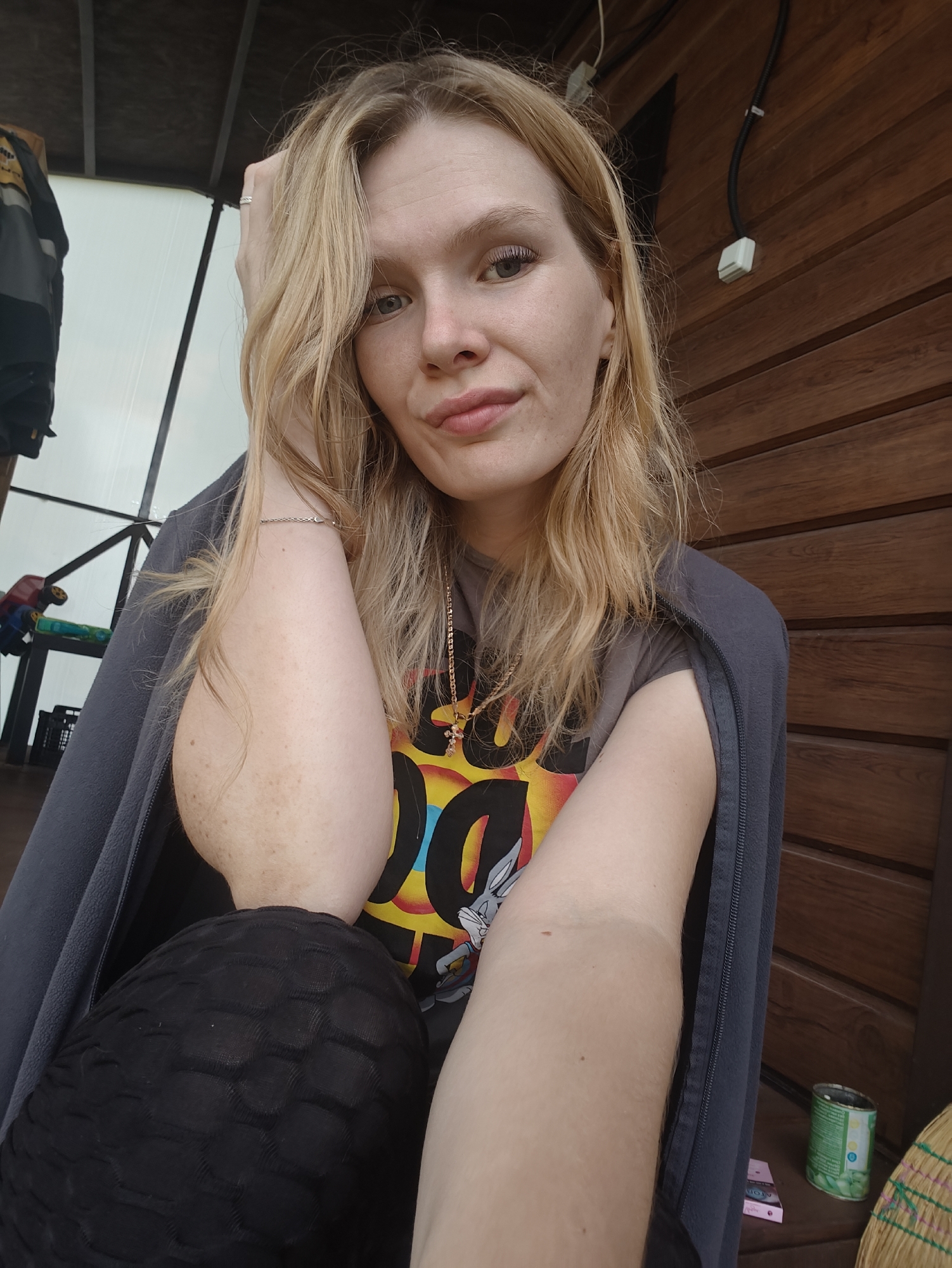 The image shows a young Caucasian woman with blonde hair, resting her head on her hand. She is wearing a gray jacket over a t-shirt with a cartoon character print. Her left arm is visible, showing freckles and a bracelet. Black textured leggings are visible on her lower body. The background consists of a wooden wall and a partially visible structure that appears to be a gazebo or porch. To the left, a jacket and some toys are visible. On the right, a can of what appears to be green beans and a broom are on a wooden surface. The scene suggests an outdoor setting, possibly in a backyard or recreational area. The image does not provide specific clues about its location within Русский Вожой, Russia.